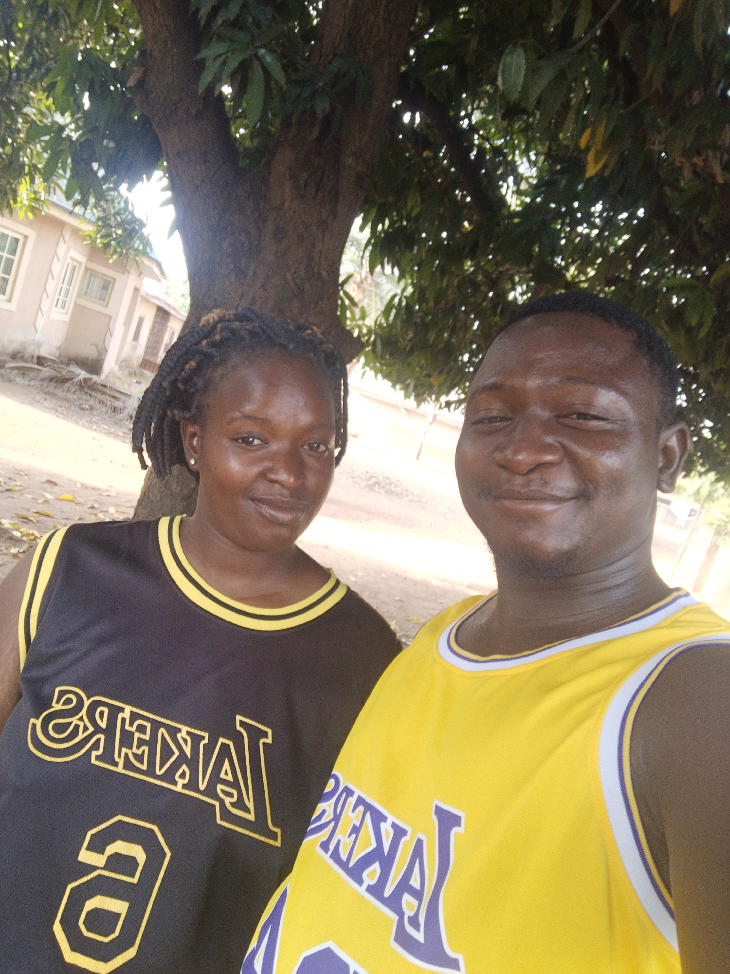 A photograph captures a man and a woman outdoors in Akwanga, Nigeria. They stand beneath a tree, its trunk visible behind them. The woman on the left is wearing a black Lakers jersey with the number 9, while the man on the right sports a yellow Lakers jersey. In the background, a building with windows can be seen. The ground appears to be dirt.