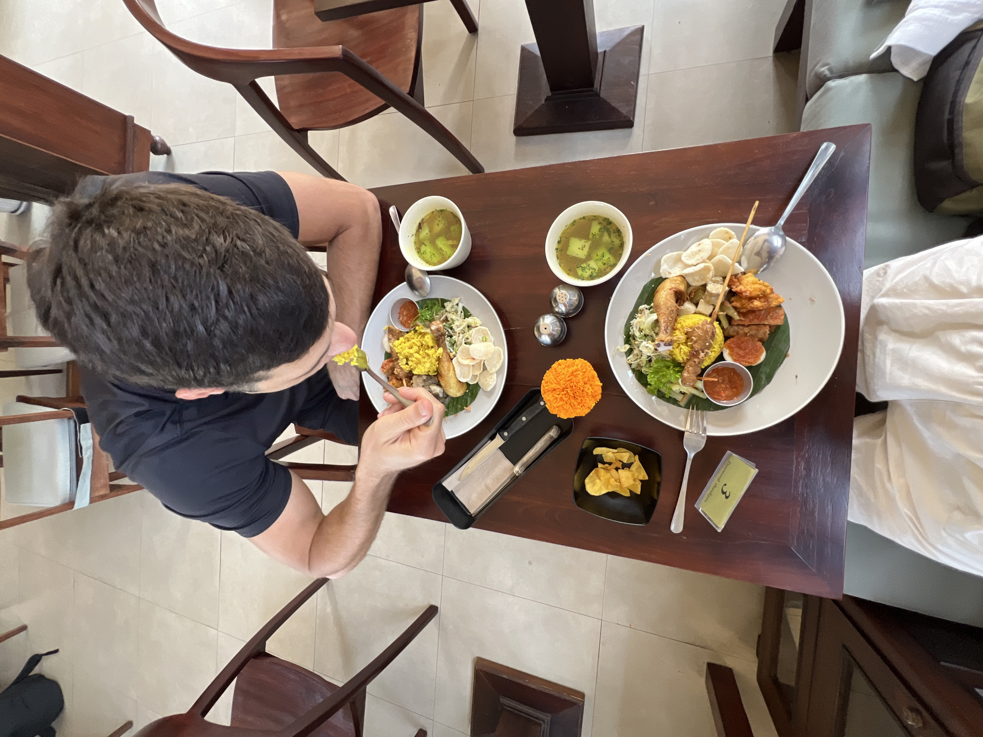 An overhead shot shows a man eating a meal at a wooden table in a restaurant in Ubud, Indonesia. The man, wearing a black t-shirt, is seated and leaning over a plate of food. The table is dark wood and is set with two plates of food, two bowls of soup, and cutlery. One plate contains rice, fried chicken, and vegetables. The other plate contains similar items. There are also small dishes of chips, a marigold flower, and a salt and pepper shaker. The floor is tiled and there are wooden chairs around the table. The scene appears to be a restaurant setting in Ubud, Bali, Indonesia.