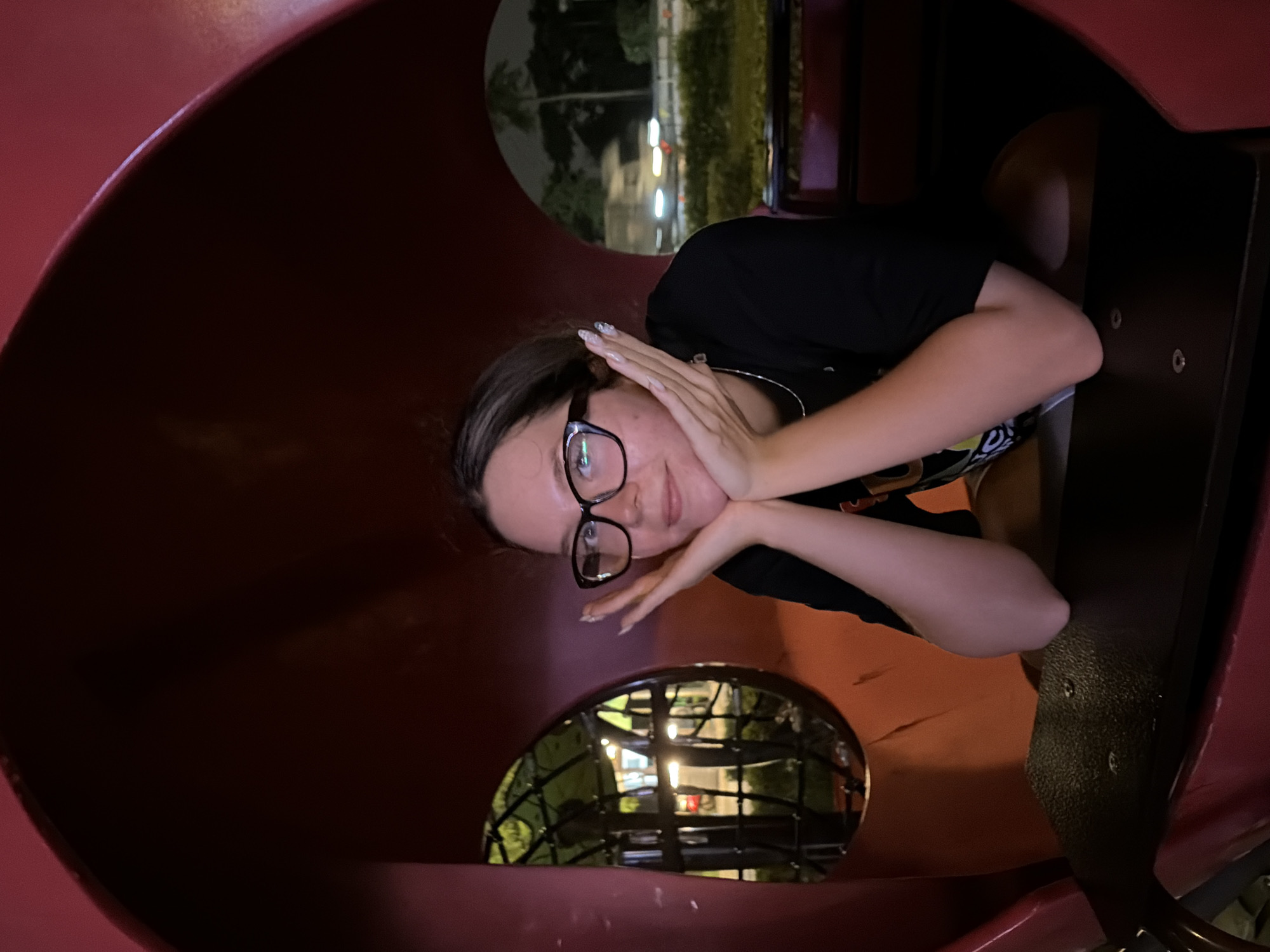 A young woman with dark hair and glasses poses for a photo, resting her chin on her hands. She wears a black t-shirt and a silver necklace. She is seated inside a red structure that appears to be part of a playground or amusement ride. Two oval-shaped openings reveal a nighttime view of trees, lights, and a blurred vehicle, suggesting an urban outdoor setting. The overall impression is that of a playful moment captured at an amusement park or similar attraction in Hanoi, Vietnam.
