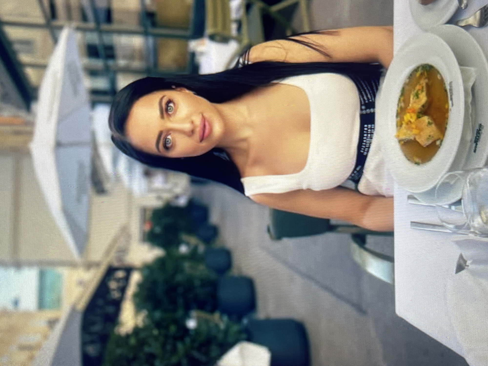 A woman with long, dark hair and light eyes is seated at a table outdoors, with a white tablecloth. She is wearing a white tank top with a black trim. In front of her is a white plate with a bowl of soup. The background is blurred, showing outdoor seating with umbrellas and greenery. The scene appears to be a cafe or restaurant in a city. However, there is no information to confirm the location as Vratsa, Bulgaria.