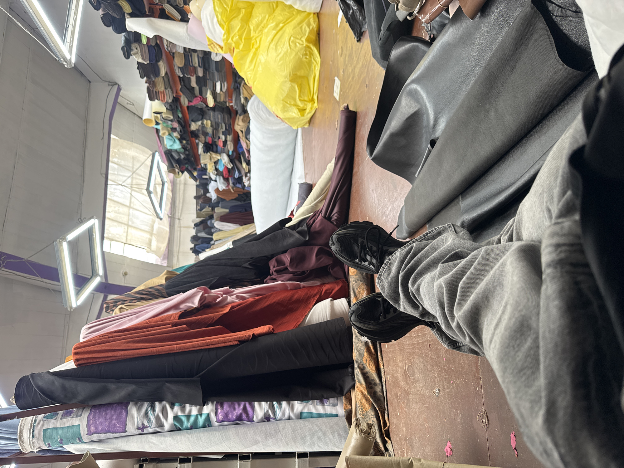 A first-person perspective shows a person sitting on the floor in a fabric store, with their legs stretched out in front of them. The person is wearing grey jeans and black sneakers. To the left, large rolls of fabric are stacked vertically, with some appearing to be patterned. To the right, several large sheets of dark leather are laid out on the floor. In the background, shelves are filled with rolls of fabric. The store appears to be well-lit with fluorescent lighting and has a skylight in the ceiling. The floor is made of reddish-brown wood. The scene is a fabric warehouse, likely in the city of Sofia, Bulgaria, given the user's request.