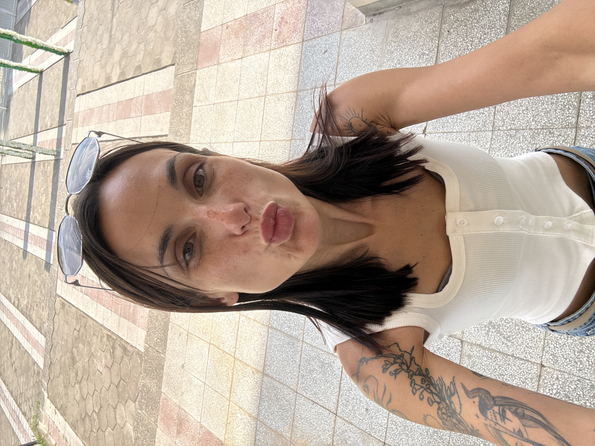 A woman with dark hair and freckles, wearing a white ribbed tank top and sunglasses perched on her head, is taking a selfie outdoors. She is puckering her lips and her left arm, which is covered in tattoos, is extended towards the camera. The background shows a paved walkway with alternating light and reddish-brown tiles. In the distance, there are decorative poles adorned with white flowers. The scene appears to be in an urban setting, possibly Pordim, Bulgaria, given the context.