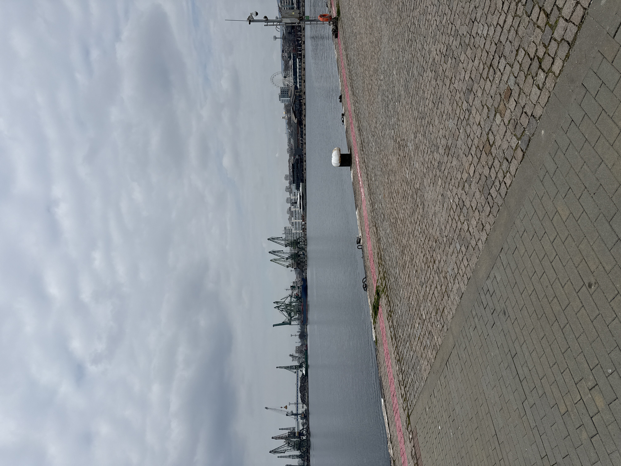 The image depicts a waterfront scene in Varna, Bulgaria. The perspective is from ground level, looking along a section of the harbor. The foreground consists of a cobblestone path and a paved area. Along the harbor's edge are bollards and markings.
In the middle ground, there is water, with cargo cranes and industrial infrastructure visible along the opposite bank. In the distance are buildings and a Ferris wheel.
The sky is overcast with a flat cloud cover.