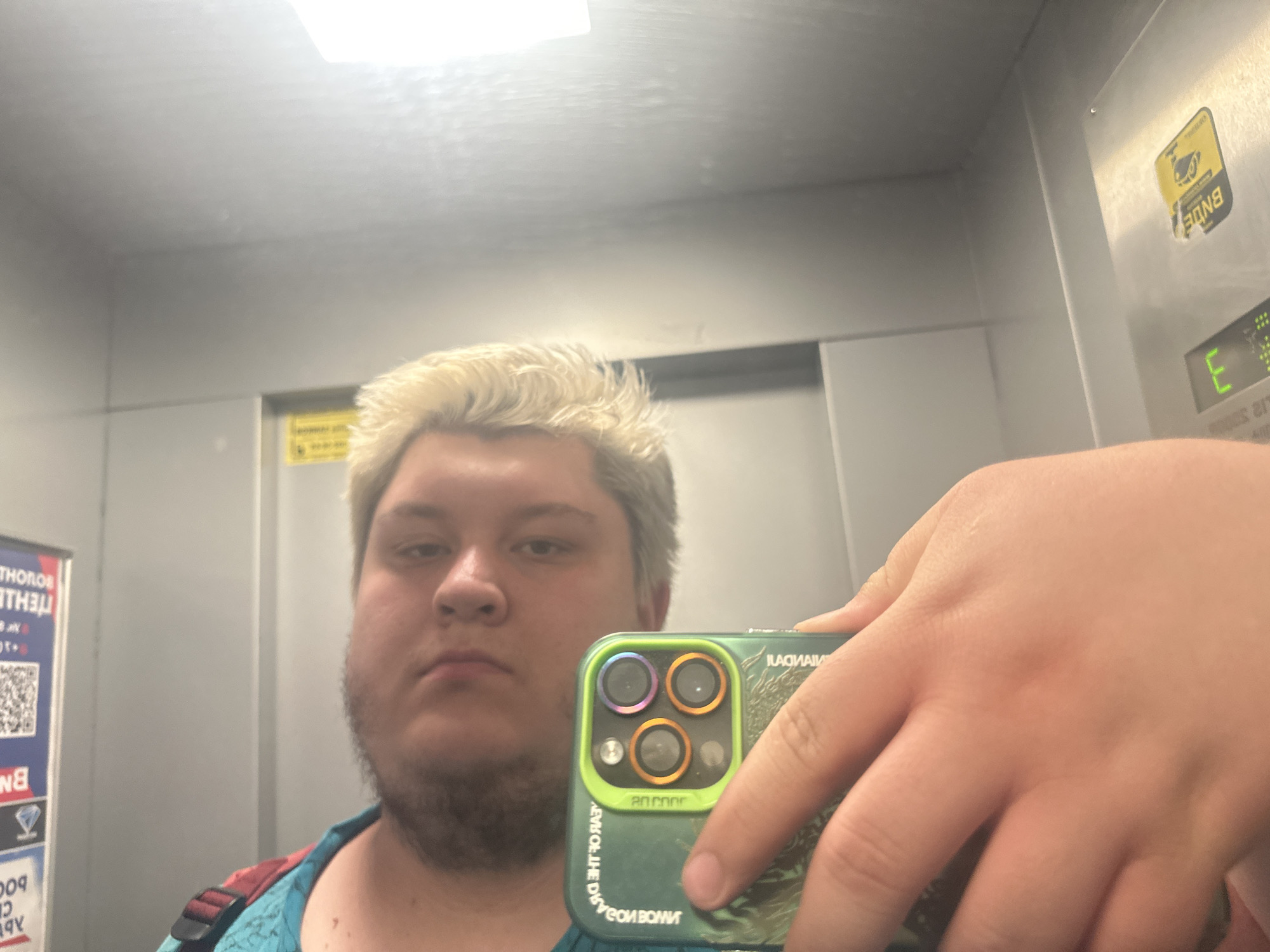 This is a selfie taken in an elevator. The subject is a person with light-colored, short, messy hair and a serious expression. They are wearing a teal shirt with a colorful strap visible over their shoulder. In their right hand, they are holding a smartphone with a green case that has a holographic effect and three camera lenses. The elevator's interior appears to be metallic and utilitarian, with a light panel on the ceiling. Visible text includes "BNDE," "E," and some text in Russian that appears to be related to elevator operations or safety. The overall impression is a casual, everyday moment captured in a somewhat confined space.