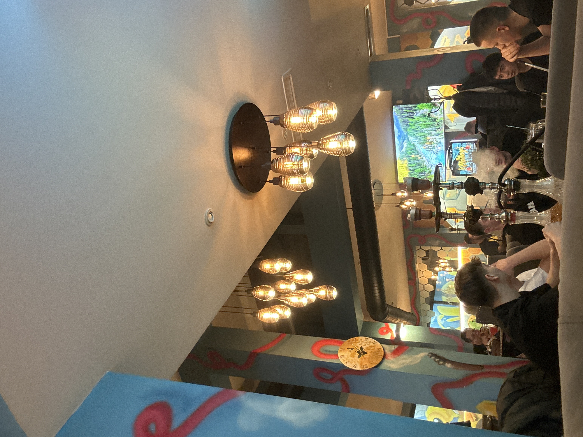 The image shows the interior of a hookah lounge. There are several people sitting and smoking hookahs. The room is decorated with colorful artwork, including cartoonish designs and graffiti-style elements. There is a television on the wall displaying a nature scene. The lighting fixtures are modern and decorative, with multiple bulbs. A clock-like wall decoration is visible. Given the overall atmosphere and details, the location may be somewhere in Sofia, Bulgaria, but this cannot be confirmed.