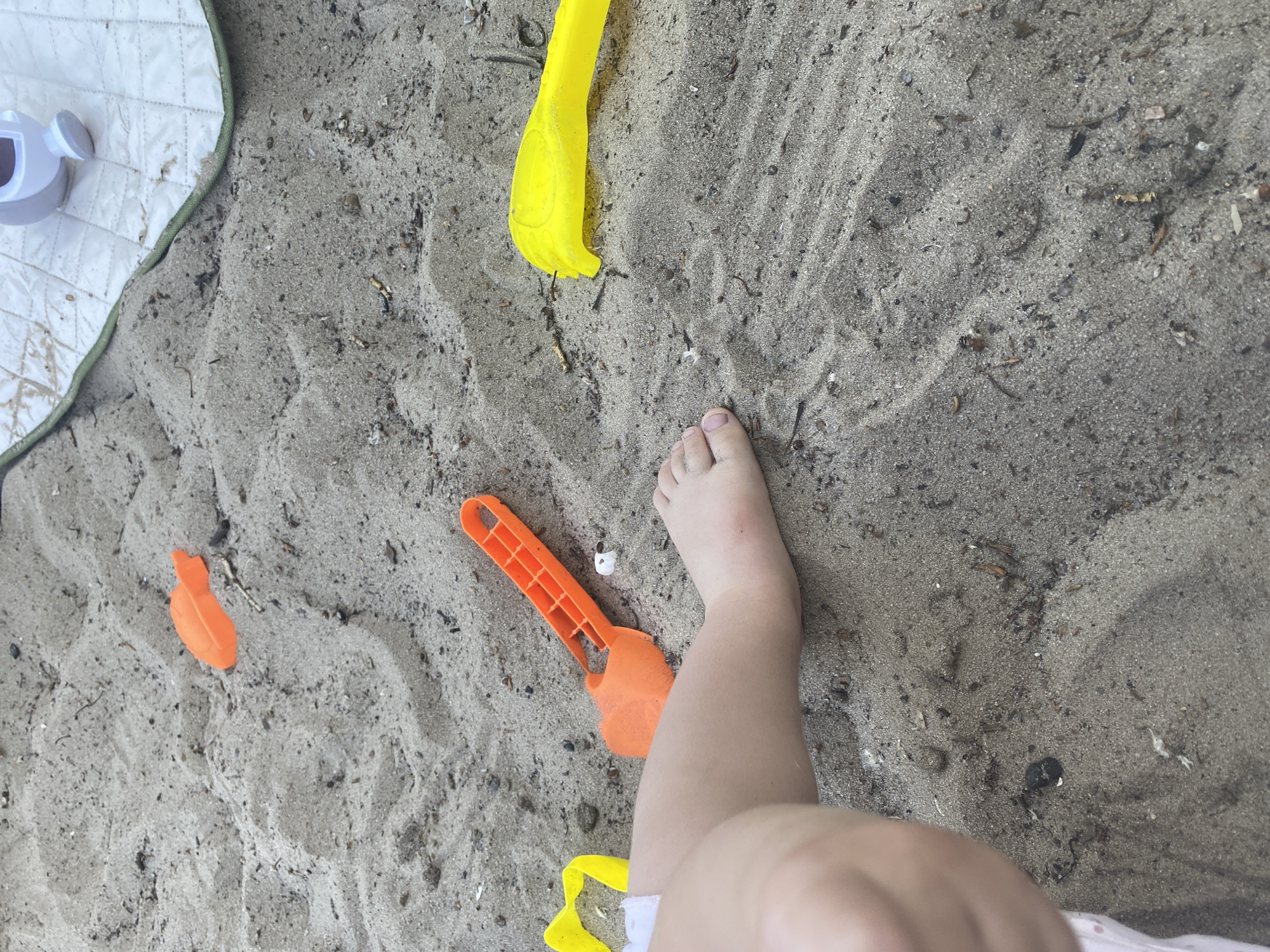 The image displays a close-up view of a sandy area, likely a beach or a sandbox, with a child's leg and foot visible in the foreground. Several colorful sand toys are scattered around: an orange shovel, a yellow shovel, and a small orange toy object. In the background, a section of a white quilted mat with green trim is visible, along with a small white toy, possibly a watering can. The sand appears wet in some areas, suggesting recent play or proximity to water. The scene is set on a cloudy day, with soft lighting casting gentle shadows. There is no discernible information within the image to indicate a specific location within the city of Dnipro, Ukraine.