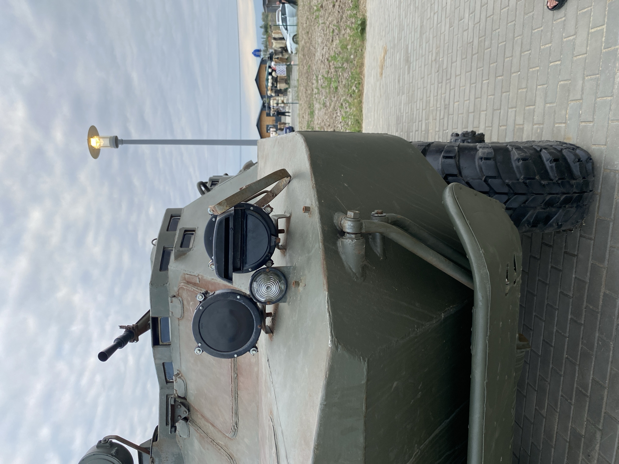 The image shows the front of a military vehicle, possibly an armored personnel carrier, parked outdoors on a paved surface. The vehicle is painted olive drab and features a prominent machine gun mounted on top. Several lights and observation ports are visible on the front. In the background, there is a lamppost, some buildings, and people milling around, suggesting a public area or event. The sky is overcast. The ground is paved with grey bricks. The vehicle is positioned at a slight angle to the camera, with its front left tire visible.