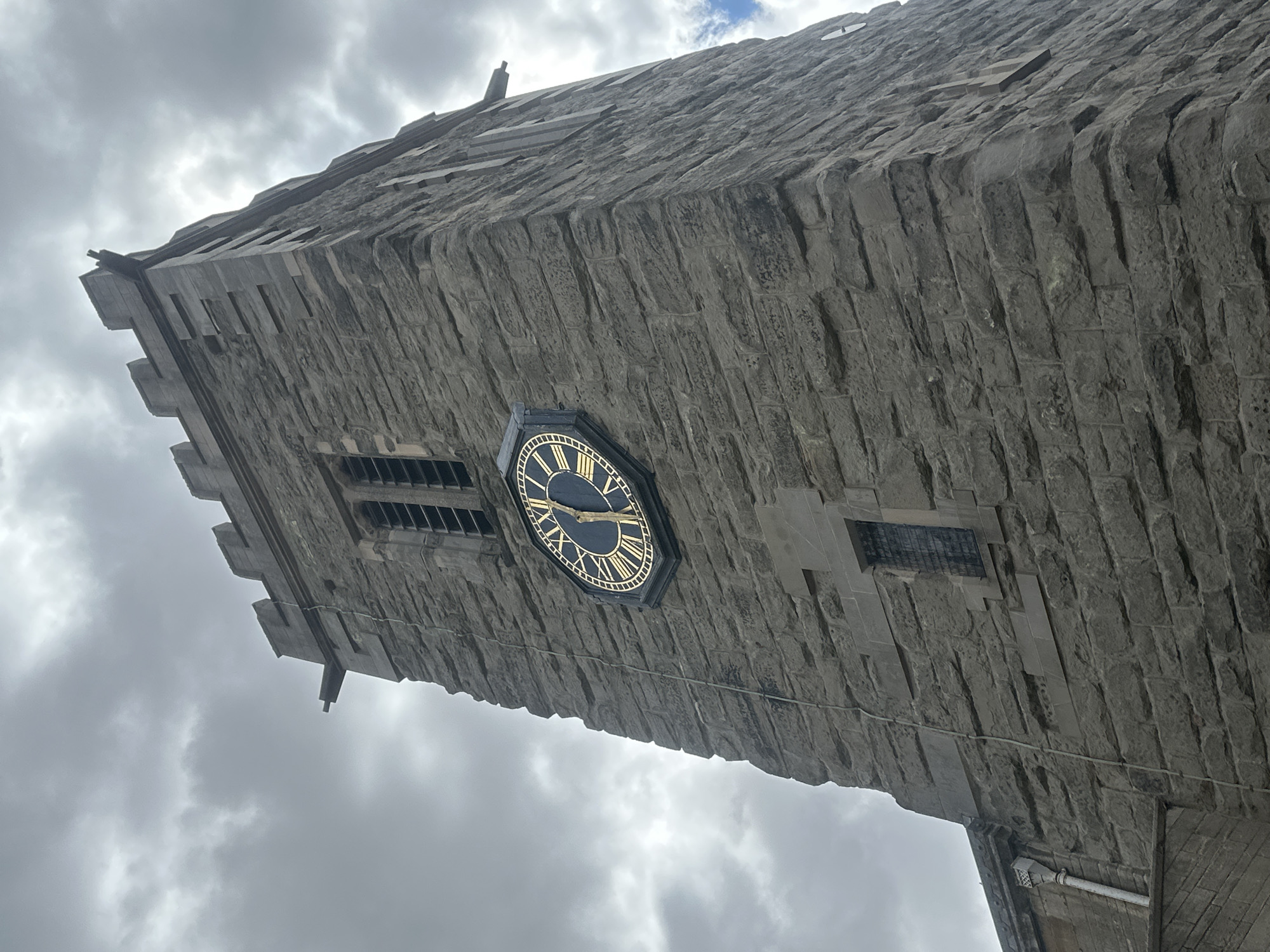 An exterior, low-angle, full shot shows a stone clock tower in Rugby, United Kingdom. The tower is made of rough-hewn grey stone blocks. At the top of the tower, there are crenellations. Gargoyles protrude from the sides of the tower, near the top. A large, circular clock face with gold Roman numerals and hands is visible on the front of the tower. The clock face has a black background and a gold border. Above the clock face, there is a narrow, rectangular window with horizontal bars. Below the clock face, there is another, smaller, narrow, rectangular window with a metal grate. A downspout runs vertically along the left side of the tower. The sky is overcast with dark, cloudy, grey clouds. The image is taken from below, looking up at the tower.