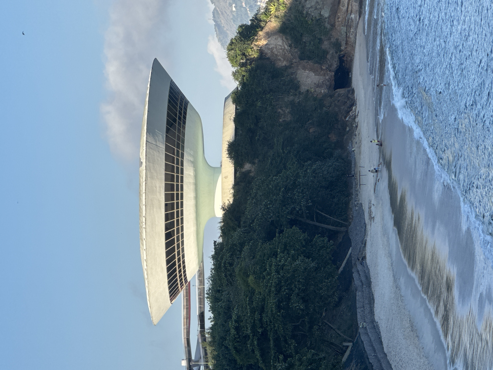 The image shows the Museum of Contemporary Art (MAC) in Niterói, Rio de Janeiro, Brazil, designed by Oscar Niemeyer. The museum, a striking saucer-shaped building, sits atop a cliff overlooking the ocean. In the foreground, a sandy beach is visible with gentle waves rolling onto the shore. Several people are scattered on the beach, some walking, others sitting. The surrounding area is lush with green trees and rocky outcrops. In the distance, a hazy mountain range can be seen. The sky is clear blue with a few wispy clouds.
