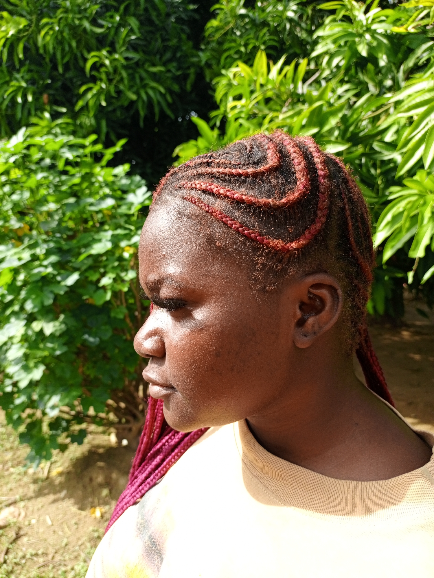 A side profile portrait captures a young Black woman outdoors in Keffi, Nigeria, on a bright, sunny day. She is positioned looking towards the left side of the frame, with her head slightly tilted. Her skin is dark-toned, and direct sunlight highlights her forehead, cheek, and shoulder.
Her hair is styled in intricate cornrows or braids, visible on her scalp in swirling, artistic patterns. Reddish-brown extensions are woven into the braids, creating a striking contrast and extending down her back, partially visible over her shoulder. She wears a simple, light-colored (likely beige or light yellow) short-sleeved t-shirt. Her left ear is visible, showing a small dark earring or piercing in the lobe. Her expression is neutral and calm.
The background consists of lush, vibrant green foliage and bushes, blurred out of focus, suggesting a natural outdoor setting like a garden or park. The strong sunlight casts shadows and creates bright spots on the leaves. There is no visible text in the image.