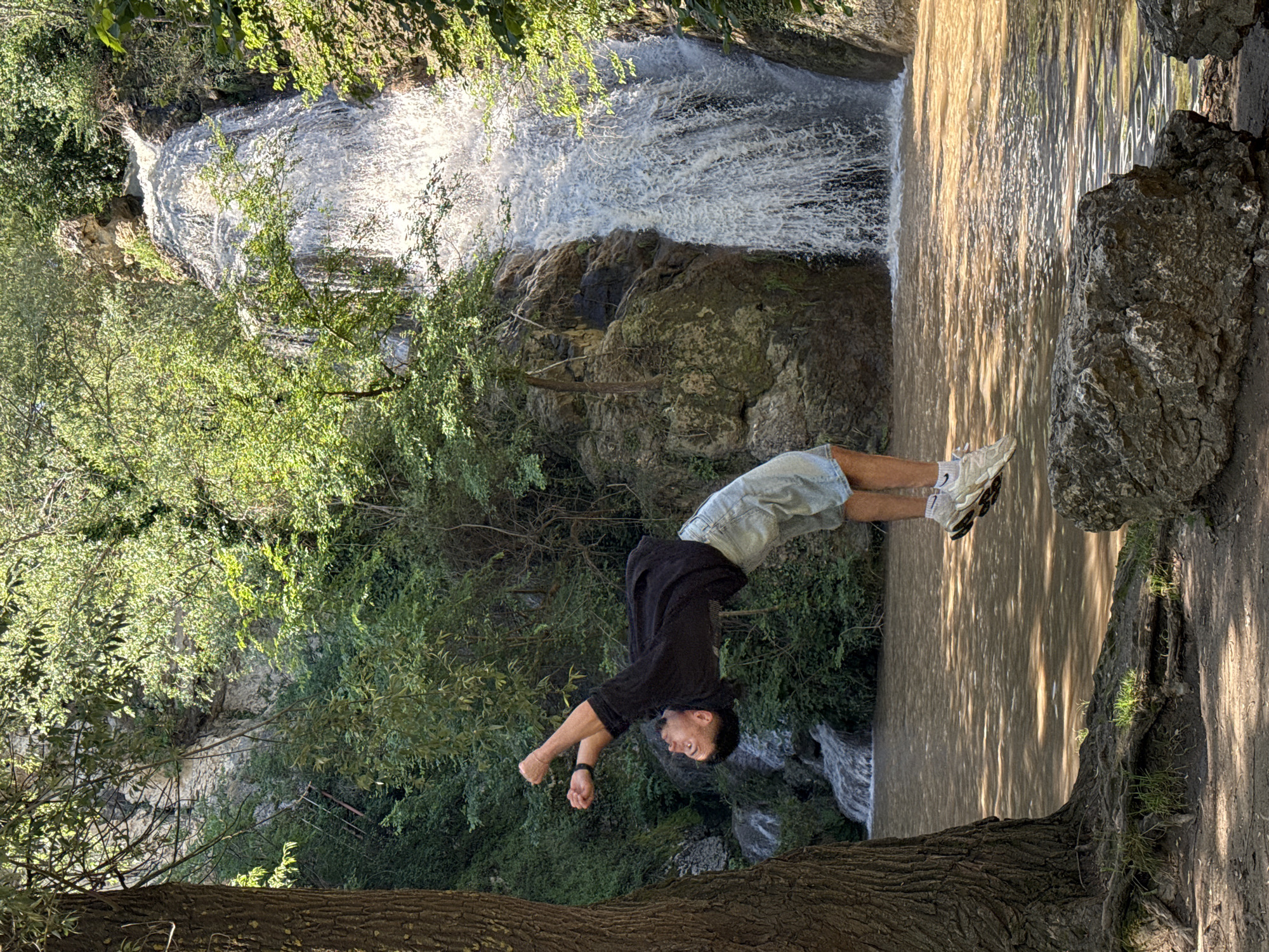 A man is performing a backflip near a waterfall in Hotnitsa, Bulgaria. He is wearing a black shirt, light blue shorts, and white sneakers. The waterfall is surrounded by trees and rocks. The water is murky. The image is rotated 90 degrees counter-clockwise.