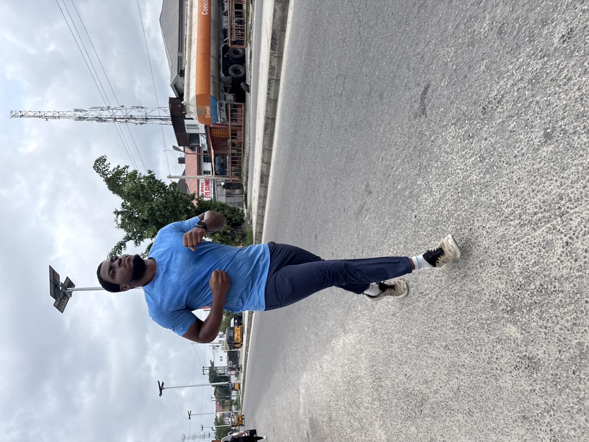 This image captures a man in motion on a paved road under an overcast sky in an urban or semi-urban setting, likely Ado, Nigeria.
The primary subject is a dark-skinned man with a beard, dressed in a light blue short-sleeved t-shirt, dark blue athletic trousers, and light-colored athletic shoes with white socks. He is mid-stride, appearing to be walking or jogging, with his arms bent at the elbows and a smartwatch visible on his left wrist. His expression is neutral and focused, looking slightly upwards and to his left.
The scene unfolds along a road, featuring a concrete curb or sidewalk to the man's right. The road surface is textured asphalt or concrete. In the background, a bustling street scene is visible. On the far side of the road, a large orange fuel tanker truck, branded "Conoil," is prominent. Behind it, various commercial buildings line the street, including one with a white sign displaying "ADAM & MICHAELS EYE CLINIC." Green trees and bushes are interspersed among the buildings.
Above the buildings and trees, a tall, slender metal tower, resembling a telecommunications or power transmission mast, rises against the cloudy sky. Several street light poles, some equipped with solar panels, are also visible. Other vehicles, including yellow "keke napep" (motorized tricycles), can be seen further down the road, indicating a typical daytime street environment in Nigeria. The weather appears to be overcast but bright, suggesting daytime conditions.