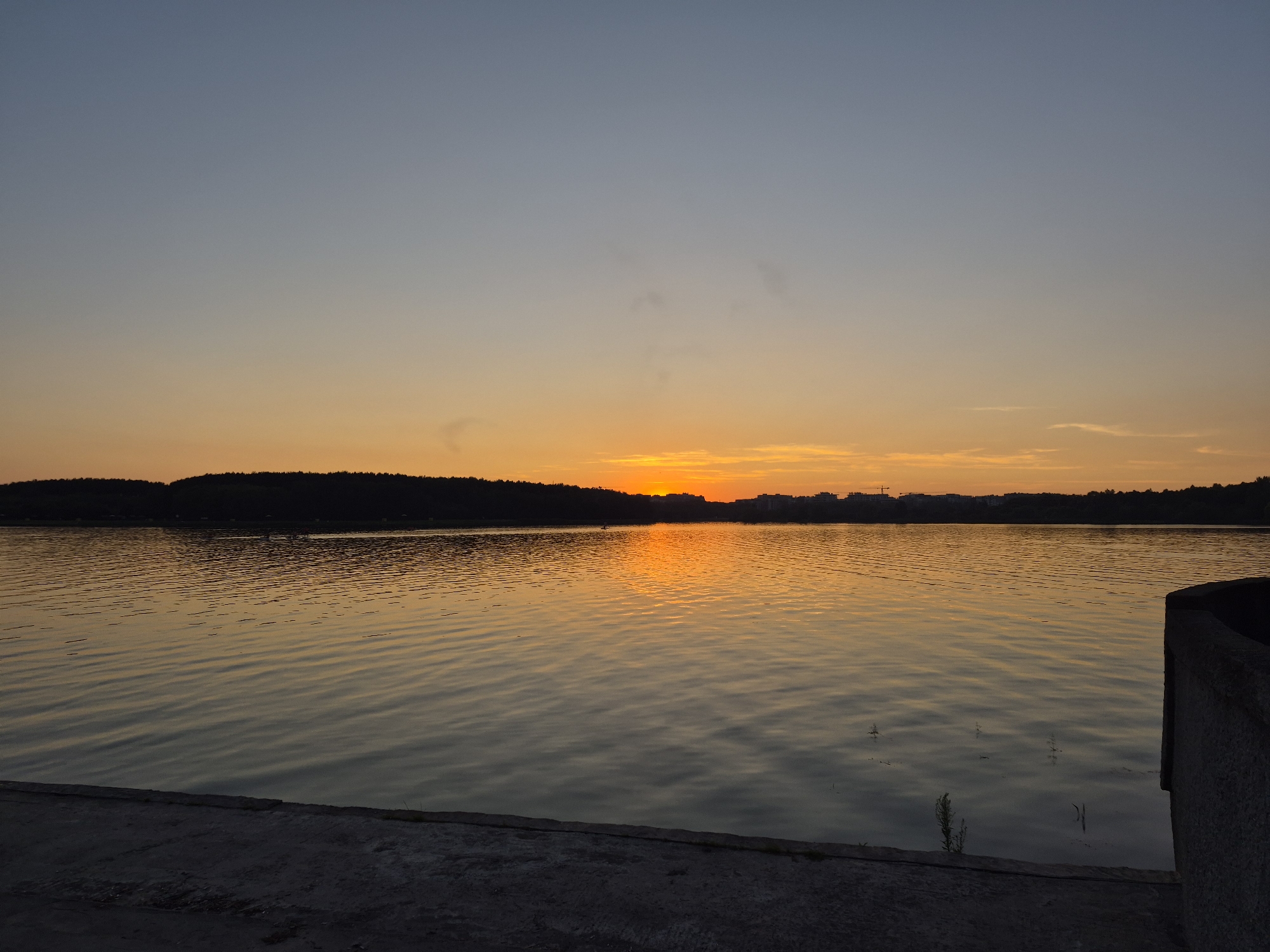 This is a horizontal, eye-level, wide shot of a lake at sunset. The sky is a gradient of colors, from a pale blue at the top to a vibrant orange and yellow near the horizon. The sun is setting behind a line of trees and buildings on the far side of the lake, casting a golden reflection on the water. The water is calm, with gentle ripples on the surface. In the foreground, there is a concrete embankment. The scene is serene and tranquil, with no people visible. This is likely the Zaslavl reservoir, also known as Minsk Sea, located near the city of Minsk, Belarus.