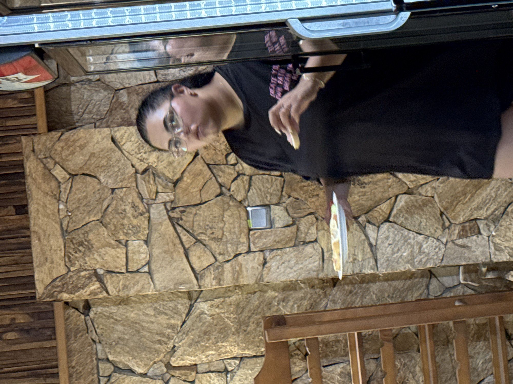 A young woman wearing glasses and a black t-shirt stands near a stone wall. She holds a white plate with food in her left hand and a piece of food in her right hand, which she is about to eat. A wooden chair is visible to her left. The background consists of a stone wall and wooden paneling. The scene appears to be indoors. The image does not provide any information about the location in Sredets, Bulgaria.