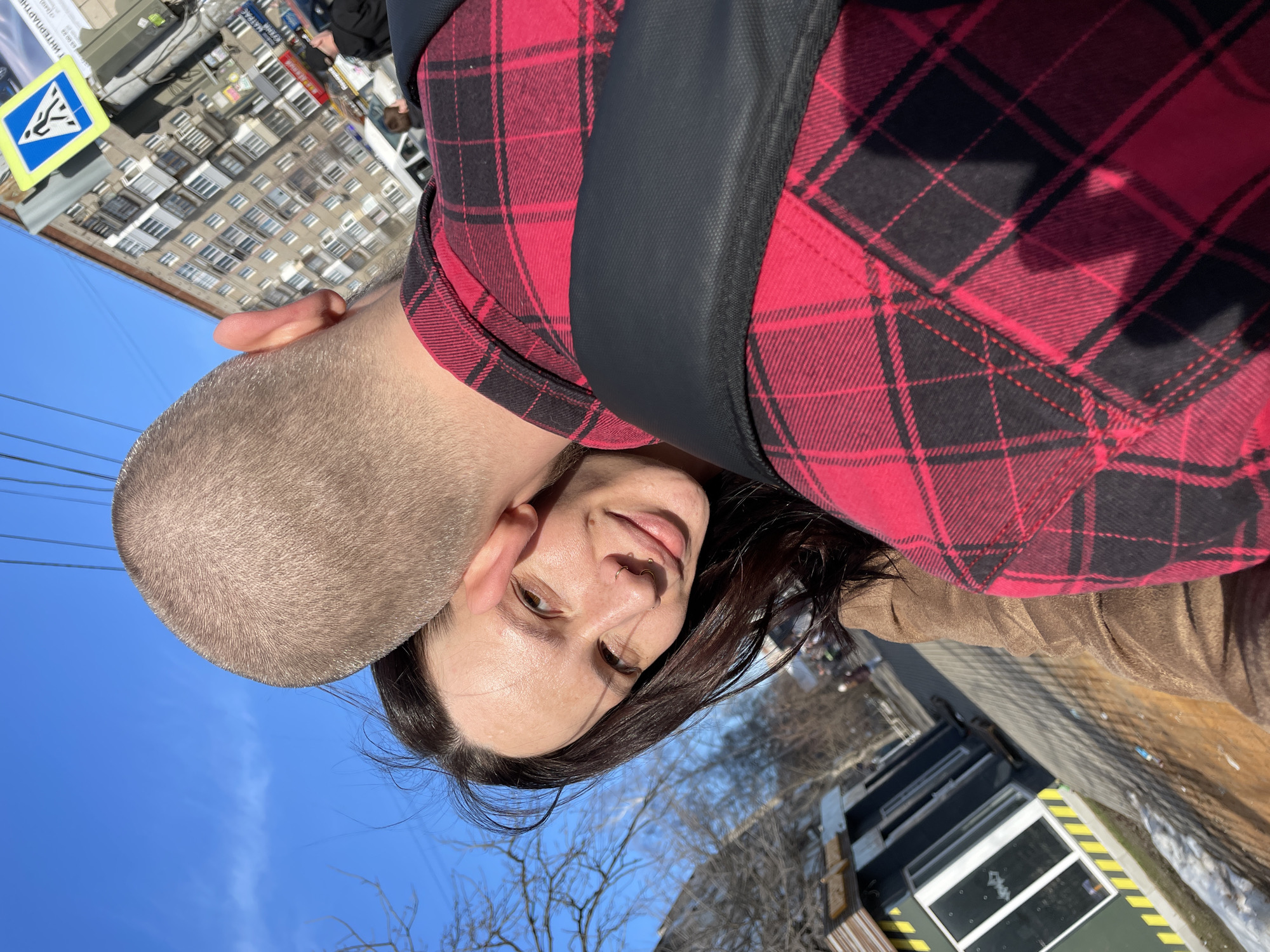 A selfie taken outdoors in Izhevsk, Russia, showing a woman with long dark hair and a nose ring, and a man with a shaved head. The woman is wearing a brown jacket and a red and black plaid shirt. The man is also wearing the red and black plaid shirt. They are close together, possibly embracing or hugging. A traffic sign and multi-story buildings are visible in the background. The sky is blue.