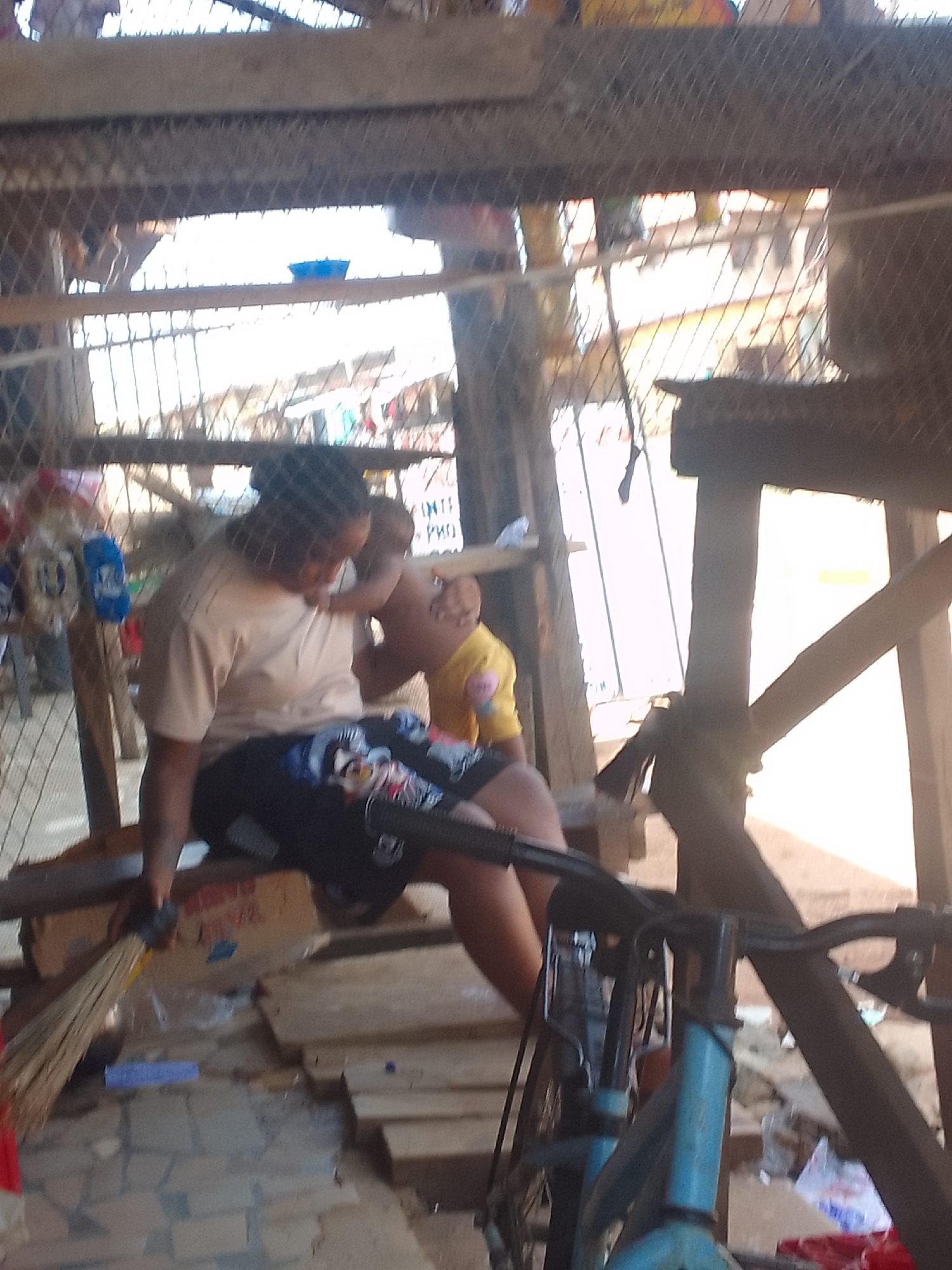 The image shows a woman and a child sitting on a wooden structure. The woman is wearing a light-colored shirt and patterned dark shorts. The child is wearing a yellow shirt. A blue bicycle is in the foreground, slightly out of focus. A broom leans against the side of the structure near the woman. The structure appears to be under a larger, possibly elevated, platform with a wire mesh covering. The environment looks like a market area, cluttered with various objects and debris. The location is Unknown, Unknown.