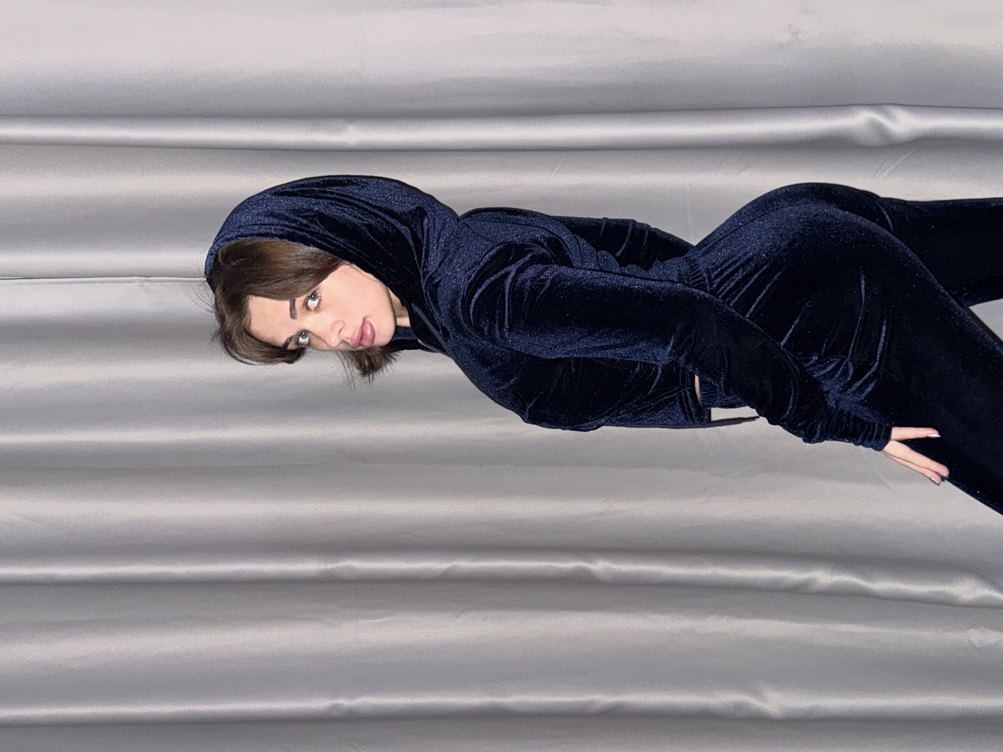 The image shows a woman lying on a silver-grey satin surface. She is wearing a dark blue velvet hooded tracksuit. The location in the city of Лобаново, Russia, is not evident from the image.