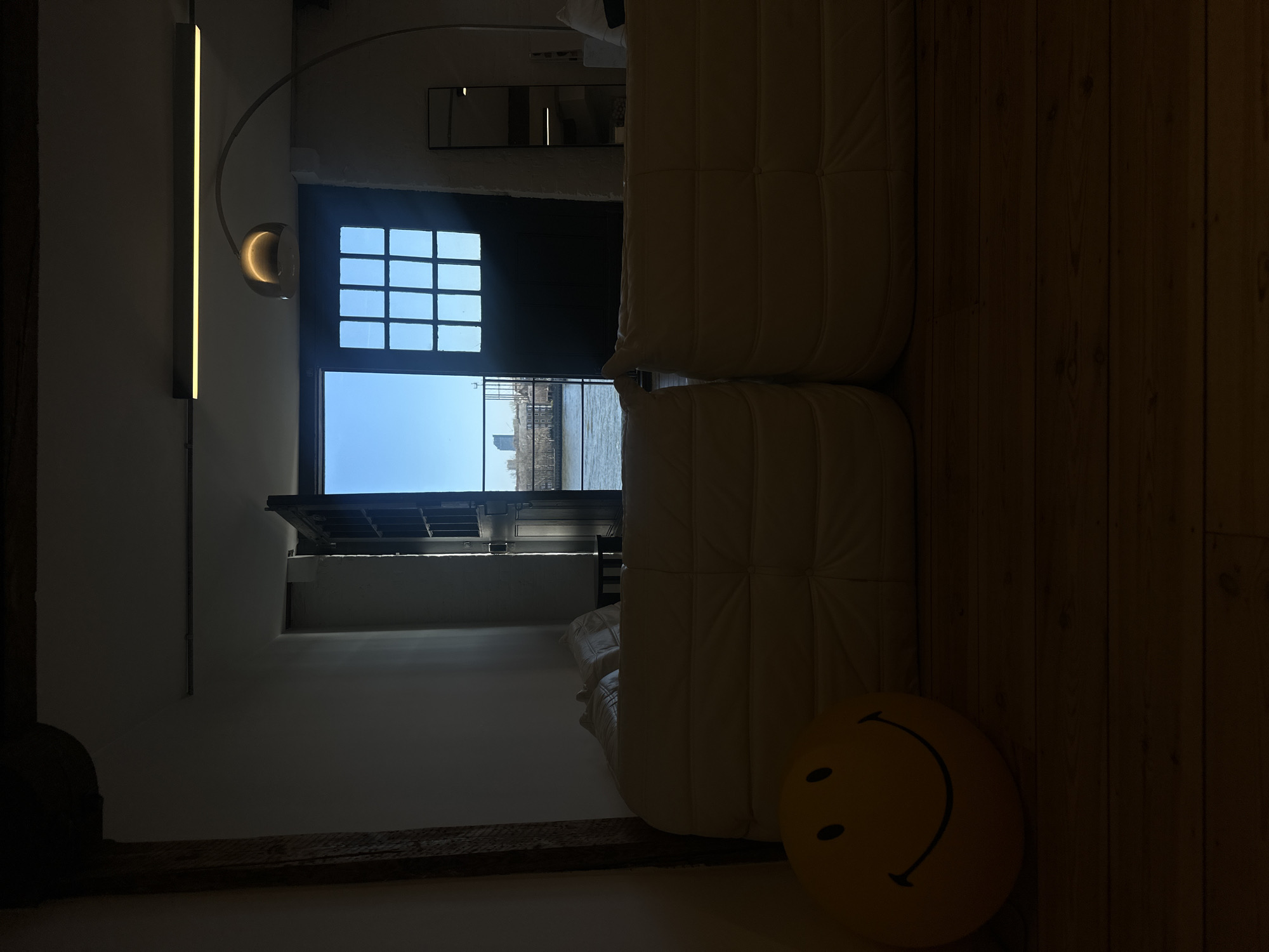 The image depicts an interior space, likely a riverside apartment or loft in the City of London, United Kingdom, looking out onto the River Thames. In the foreground, a cream-colored, multi-segment modular sofa occupies the lower half of the frame. To the left of the sofa, a large yellow spherical pillow featuring a black smiley face rests on dark wooden floorboards. A dark wooden structural beam runs vertically along the left edge of the room. The interior walls are white.
In the mid-ground, a dark, double-doored opening, with multiple glass panes forming a grid in its upper section, reveals the exterior. Through this opening, a wide body of water, identifiable as the River Thames, is visible. On the distant bank, a cityscape with various buildings and tree lines is present under a clear blue sky. A prominent, modern glass skyscraper, consistent with landmarks like The Shard, stands distinctly against the horizon on the opposite side of the river.
The interior ceiling features a long, linear, illuminated light fixture. To the right, an arched floor lamp with a spherical, illuminated head extends into the upper portion of the frame. A tall, narrow, black-framed mirror is positioned against the right wall, reflecting a part of the room. The interior lighting is dim compared to the bright exterior, suggesting natural light entering from the open doors. No individuals are present in the scene.