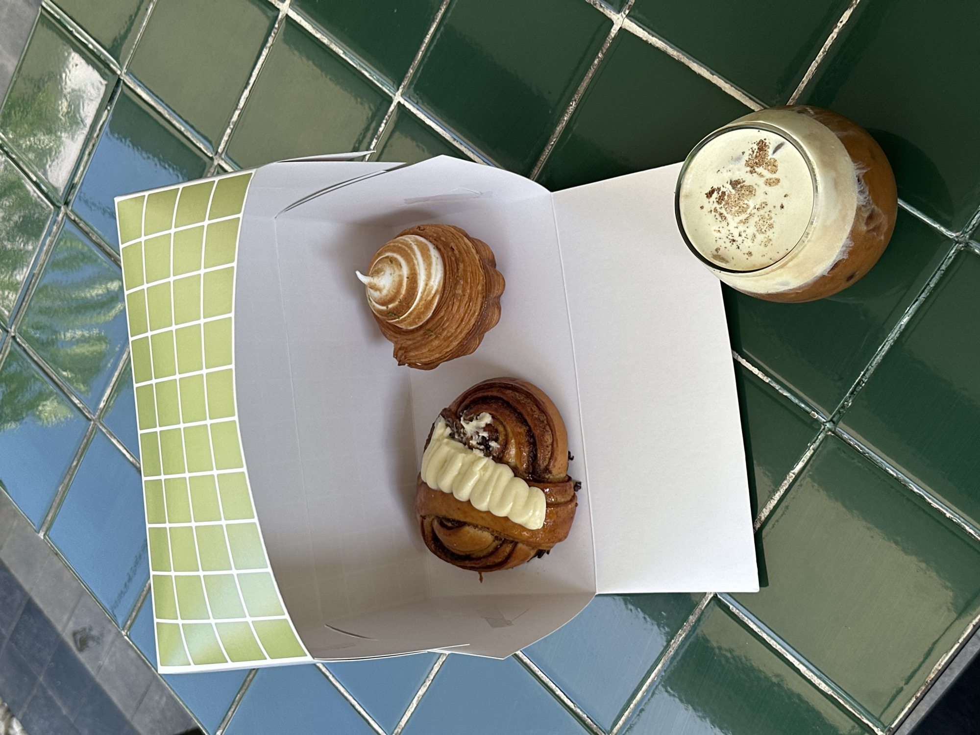 A high-angle shot shows a white box containing two pastries and a glass of iced coffee on a surface covered with green and blue square tiles. The box is white with a light green grid pattern on one side. Inside the box, there is a pastry with white meringue on top and a cinnamon roll topped with white icing. Beside the box is a glass of iced coffee with a layer of white foam and brown sprinkles on top. The environment suggests the Ubud District, Indonesia.