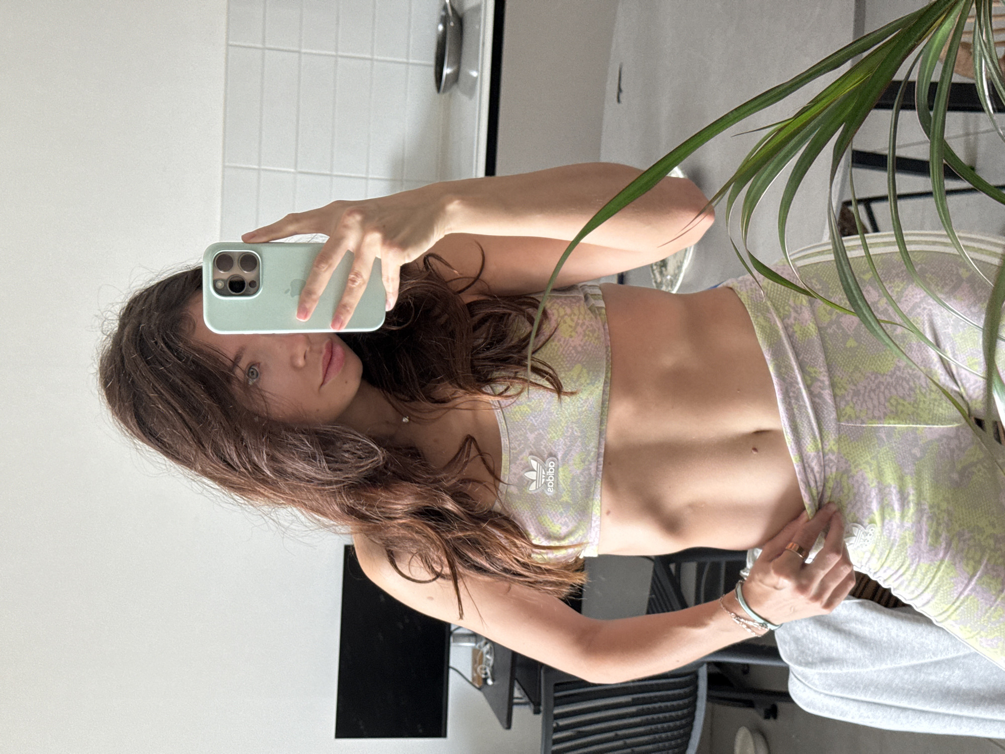 A woman is taking a selfie in what appears to be a bathroom or kitchen setting. She is wearing a patterned Adidas sports bra and matching pants. Her long brown hair is partially obscuring her face. The background features white tiled walls, a stainless steel sink, a black television, and a glimpse of indoor foliage, possibly a houseplant in a pot. The location in Givatayim, Israel, is not identifiable from the visual information provided.