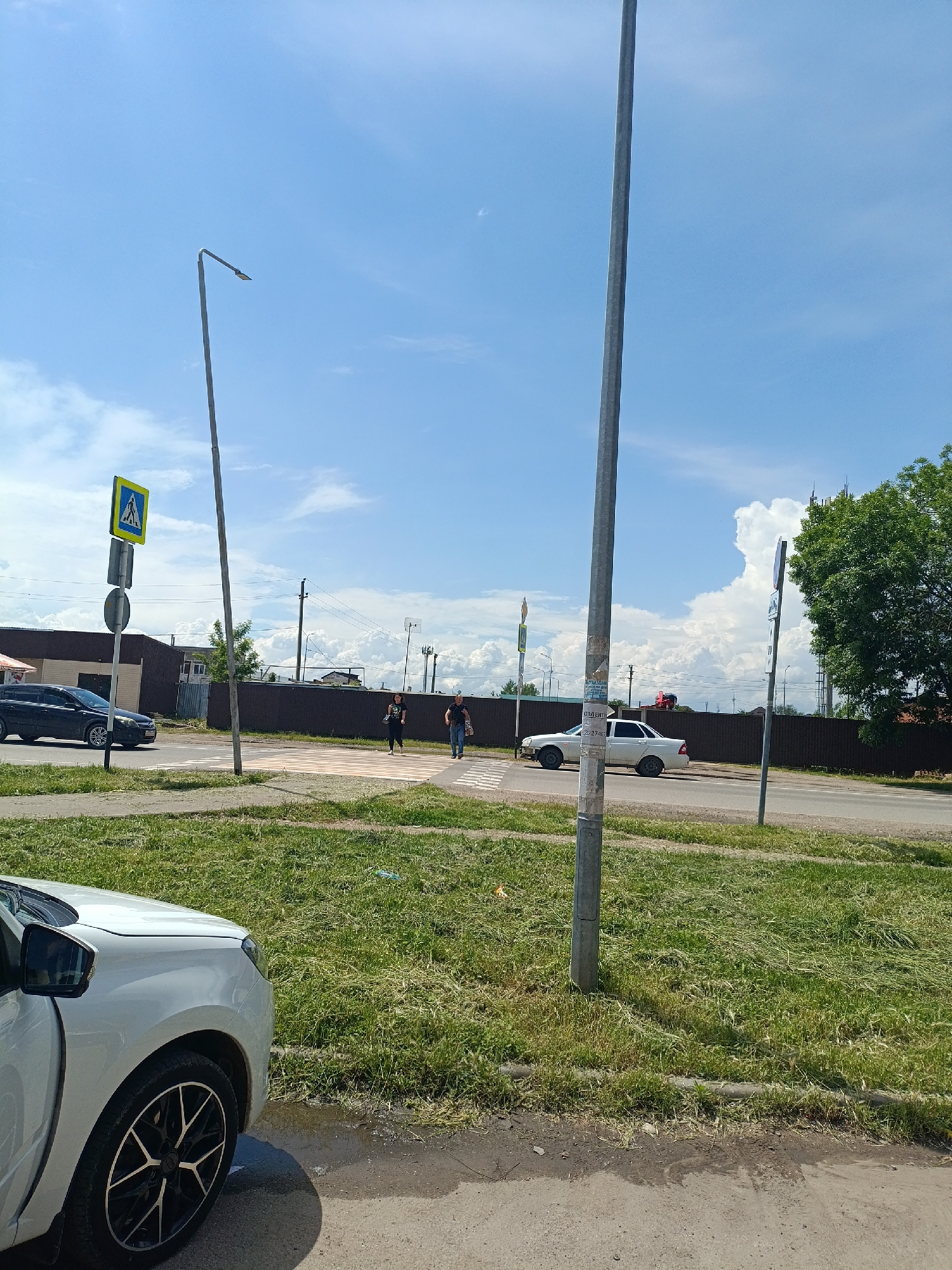 The image shows a street scene in Yablonovskiy, Russia on a sunny day with scattered clouds. A crosswalk is visible with two people crossing the street. Cars are parked on either side of the road. There are street lights and signs present. A white car is in the foreground on the left side of the image. There's a strip of grass between the sidewalk and the street. A brown fence runs along the edge of the buildings and tree on the right side of the road.