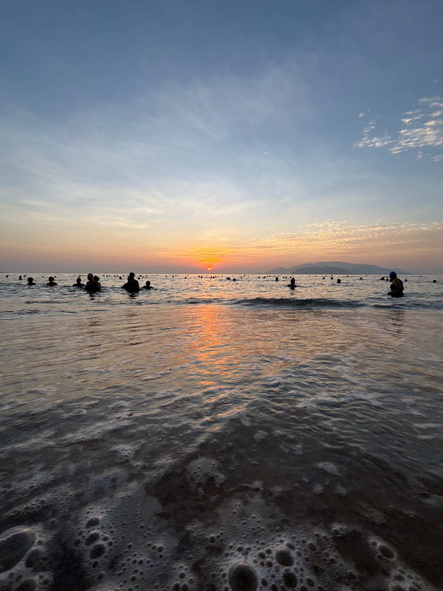 This image depicts a sunset over the ocean at a beach in Nha Trang, Vietnam. The sun is low on the horizon, casting a warm orange glow across the sky and reflecting on the water's surface. Numerous silhouetted figures of people are visible wading in the shallow water, some appearing to be swimming or enjoying the evening. The foreground shows gentle waves breaking and creating foam as they approach the shore. In the distance, a hazy silhouette of mountains can be seen against the twilight sky. The overall atmosphere is serene, characteristic of a popular beach destination at dusk.