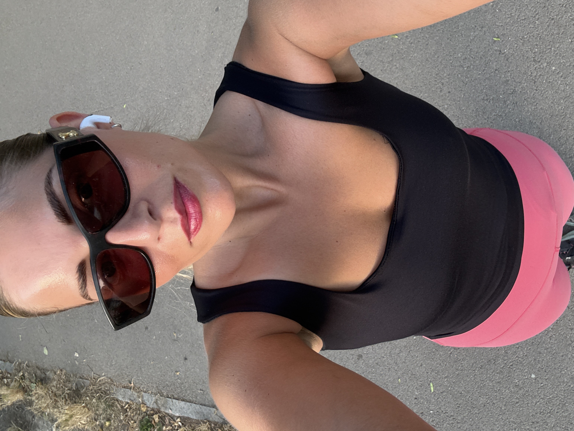 A woman is taking a selfie outdoors on a paved path. She is wearing a black tank top, bright pink leggings, black sunglasses, and white wireless earbuds. Her hair is pulled back, and she is wearing lipstick. The background shows dry grass on the left and the paved path on the right. There are no discernable landmarks or clues to indicate the location is Paris, France.
