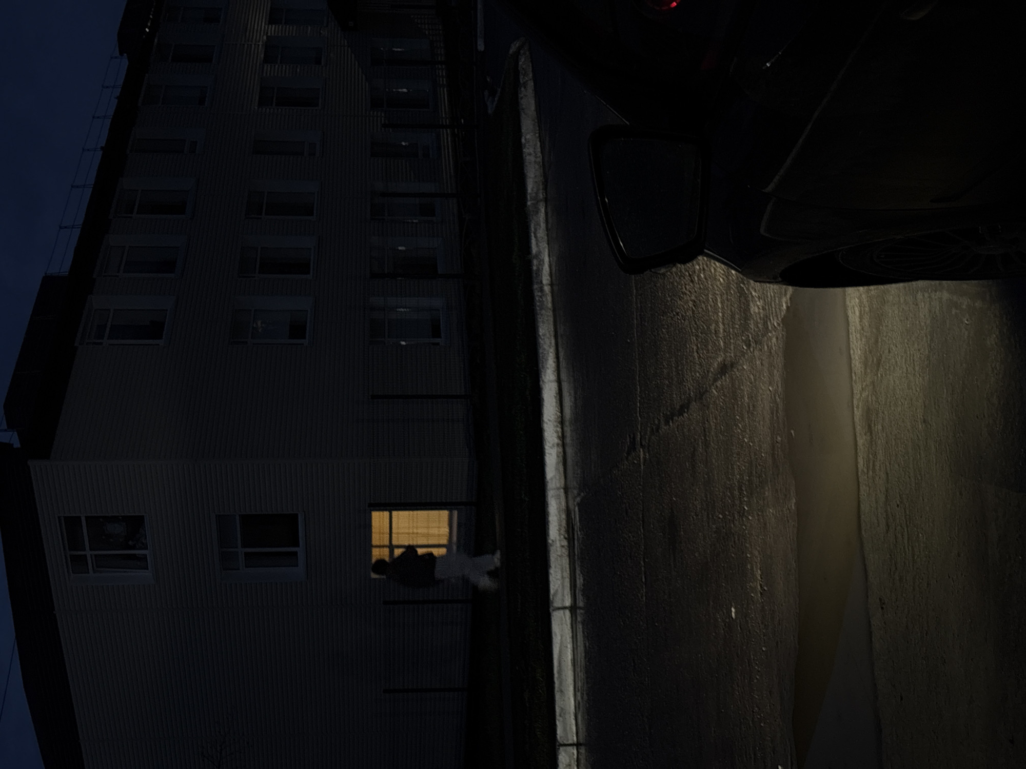 A nighttime shot taken in what appears to be Chaikovsky, Russia, shows a tall building adjacent to a narrow alley. A dark-colored vehicle, specifically the side with a wing mirror and partial view of the undercarriage, is positioned near the alleyway. Most of the windows in the building are dark, but one window is illuminated, revealing two people within. The alley is dimly lit, possibly by the vehicle's headlights, and its walls appear textured. The overall scene has a low-light and somewhat unsettling atmosphere.