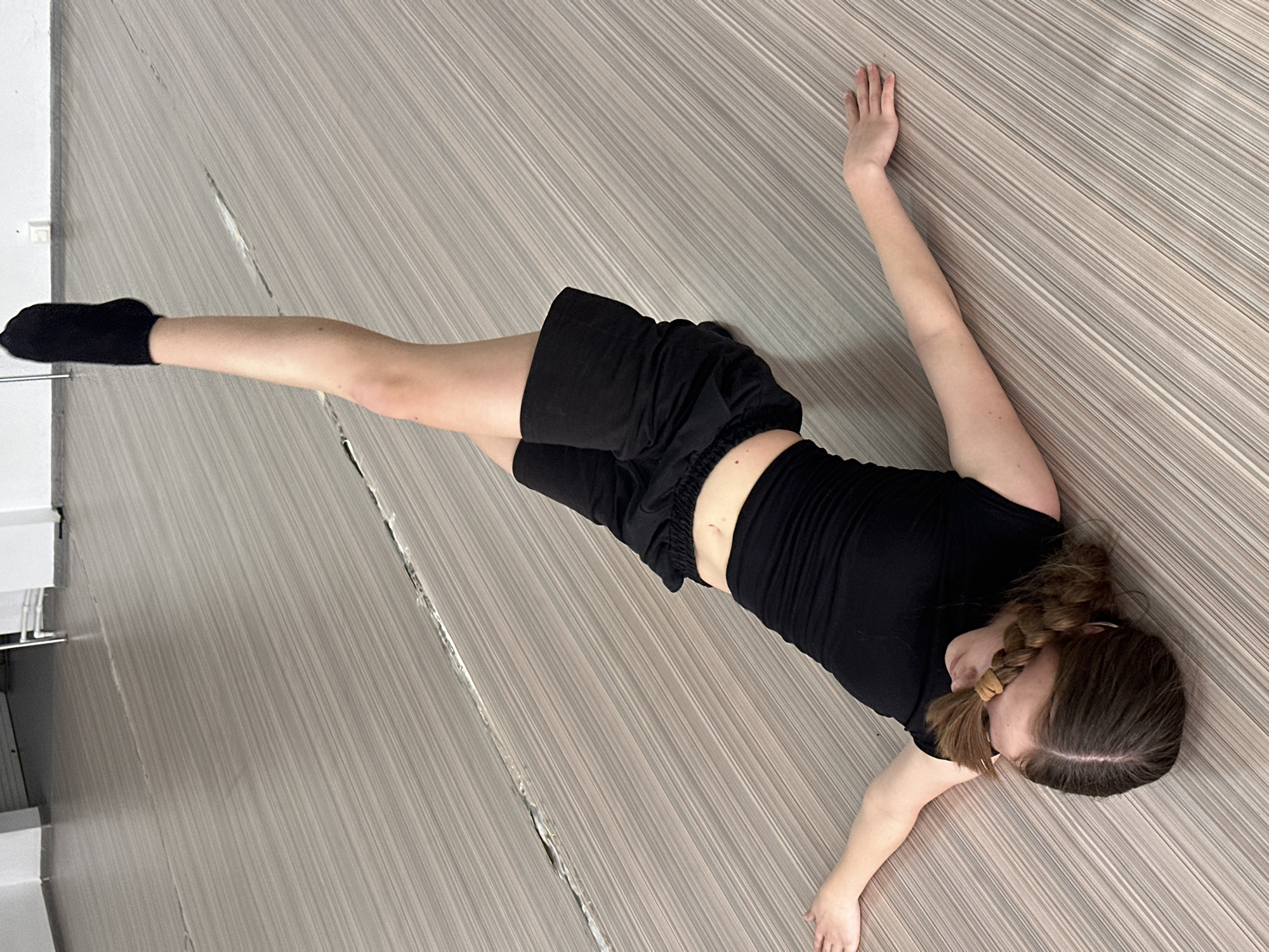 A young person wearing black shorts, a black t-shirt, and a black sock is lying on their back on a floor of gray and white striped panels. Their right leg is extended away from their body, and their arms are outstretched on either side of their head. Their hair is styled in braids. There is no visible evidence indicating the location of Votkinsk, Russia, or any other city.