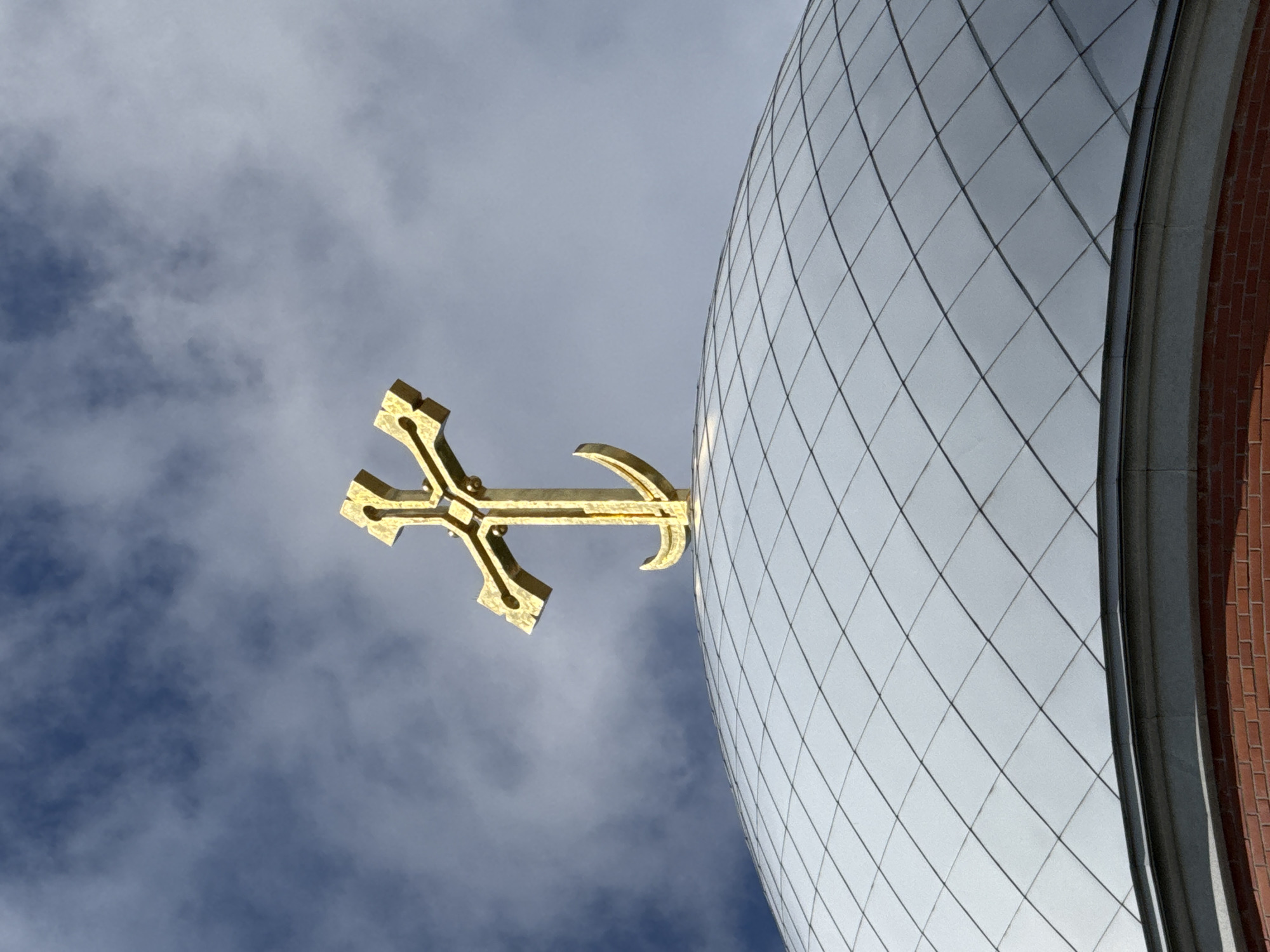 A golden Orthodox cross is mounted on a silver-tiled dome of a building in Saint Petersburg, Russia. The sky behind the cross is a mix of blue and grey clouds. The dome is made of diamond-shaped tiles, and the base of the dome is visible, constructed of red brick with a grey stone trim. The image is a close-up, focusing on the cross and the upper part of the dome.