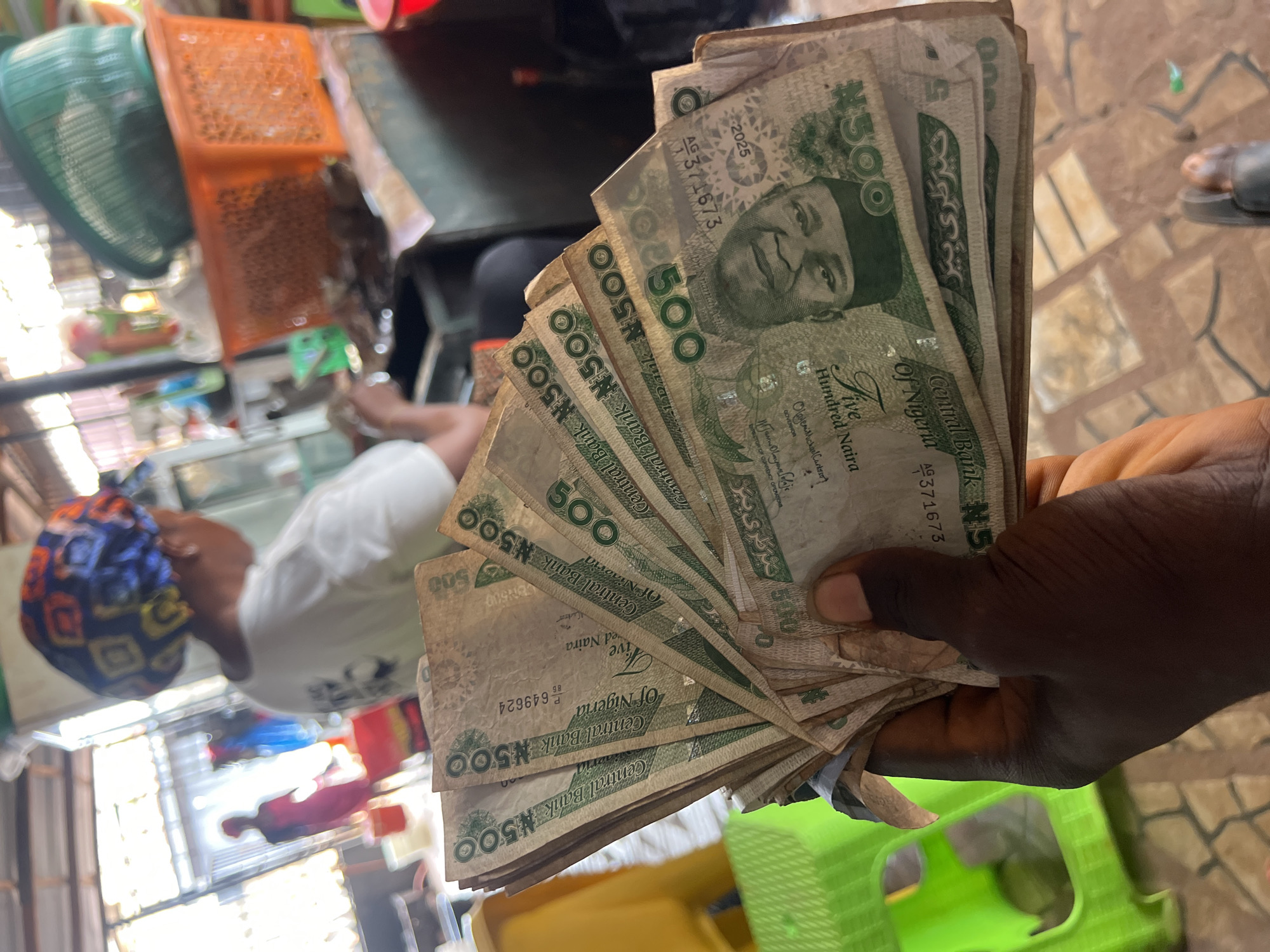 A person's hand holds a fan of Nigerian 500 Naira banknotes in an outdoor market setting. The banknotes are predominantly green and feature the portrait of Nnamdi Azikiwe. In the background, out of focus, is a woman wearing a colorful headwrap and white shirt, possibly a vendor. To the right, orange and green baskets are stacked. The ground appears to be tiled. The overall scene suggests a transaction or display of money in a busy market environment, likely within Nkpor, Nigeria, given the currency.