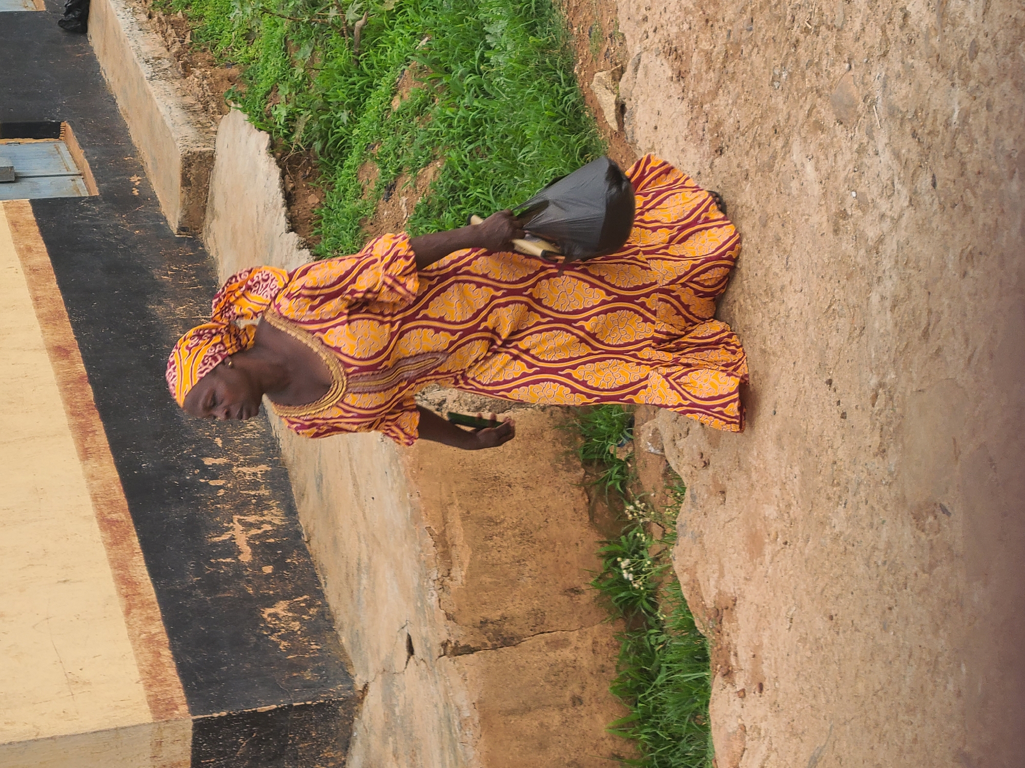 A woman in a yellow and maroon patterned dress and headwrap stands outdoors, holding a black plastic bag. The background consists of a wall with black and tan sections, patches of green grass, and a dirt ground. The location is Unknown, Unknown.