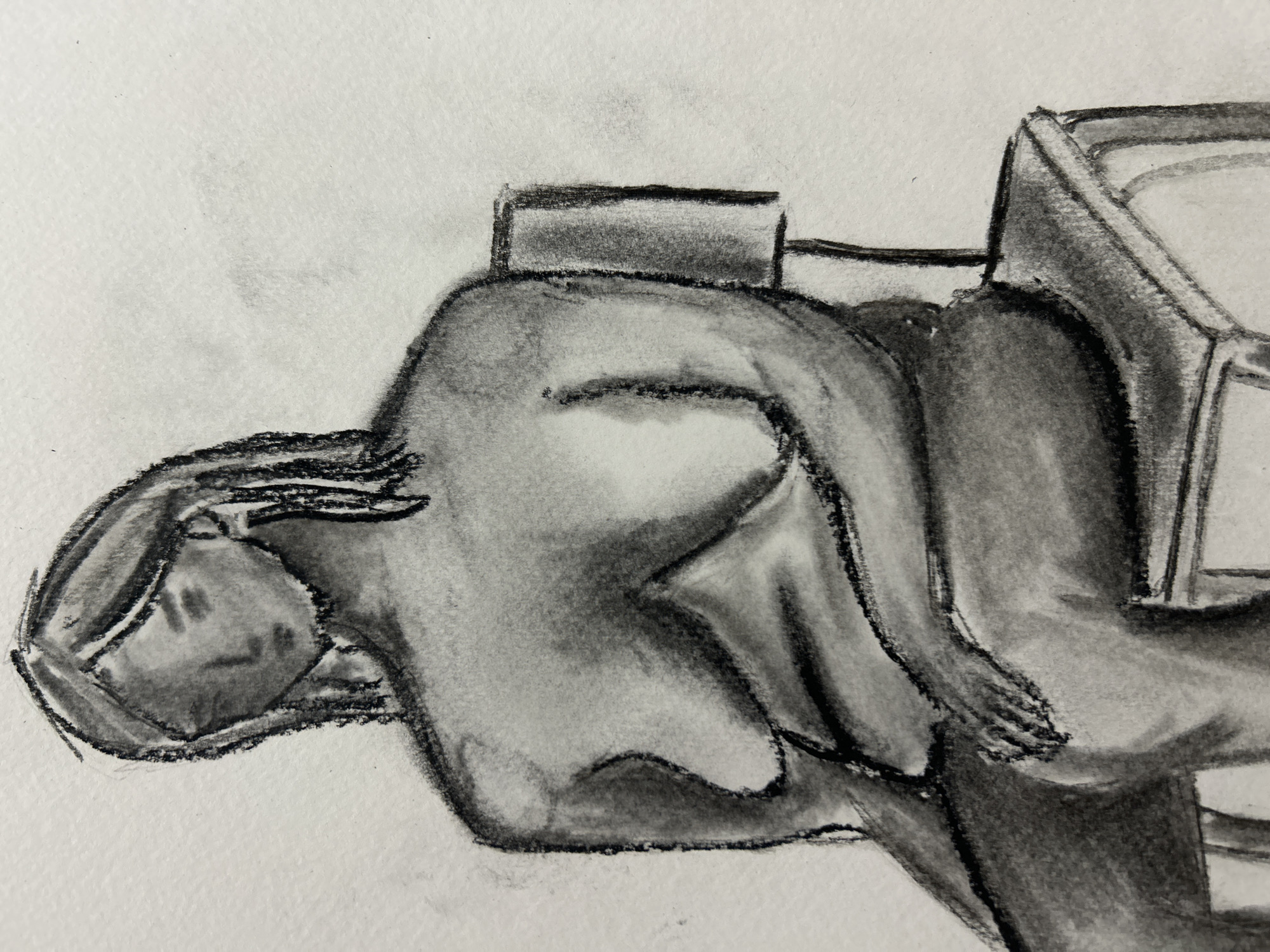 A charcoal drawing depicts a figure reclining in a chair, possibly reading. The person is partially nude, with emphasis on the torso and limbs. The chair has a boxy shape and appears to be made of solid material. The drawing is rendered in shades of gray, with varying degrees of darkness to create depth and form. The style is suggestive rather than photorealistic. There is no evidence to suggest it is located in Sofia, Bulgaria.