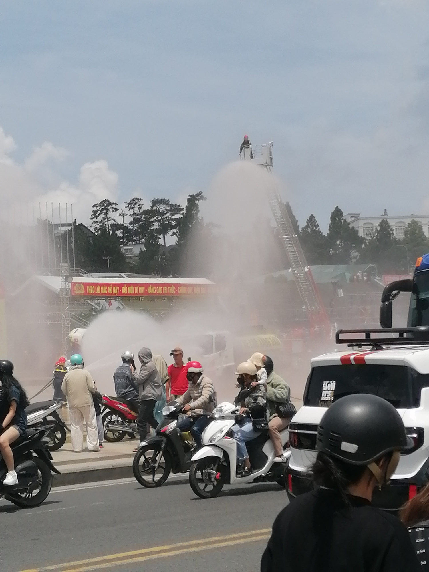 In Da Lat, Vietnam, a scene unfolds involving emergency services and onlookers. A large fire truck is present, with its ladder extended and a strong stream of water being sprayed into the air, creating a significant mist. On the ladder, a person in a red helmet and dark clothing appears to be operating the water spray. Below, on the ground, several individuals are gathered. Some are wearing helmets and appear to be on or near motorcycles, suggesting they might be spectators or involved in a demonstration. Others are dressed in everyday clothing, observing the activity. A white vehicle, possibly a van, is parked nearby, and a banner with Vietnamese text is visible in the background, indicating a public event or demonstration. The overall environment suggests an outdoor setting with some buildings and trees in the distance, under a partly cloudy sky. The event seems to be taking place on a street in Da Lat.