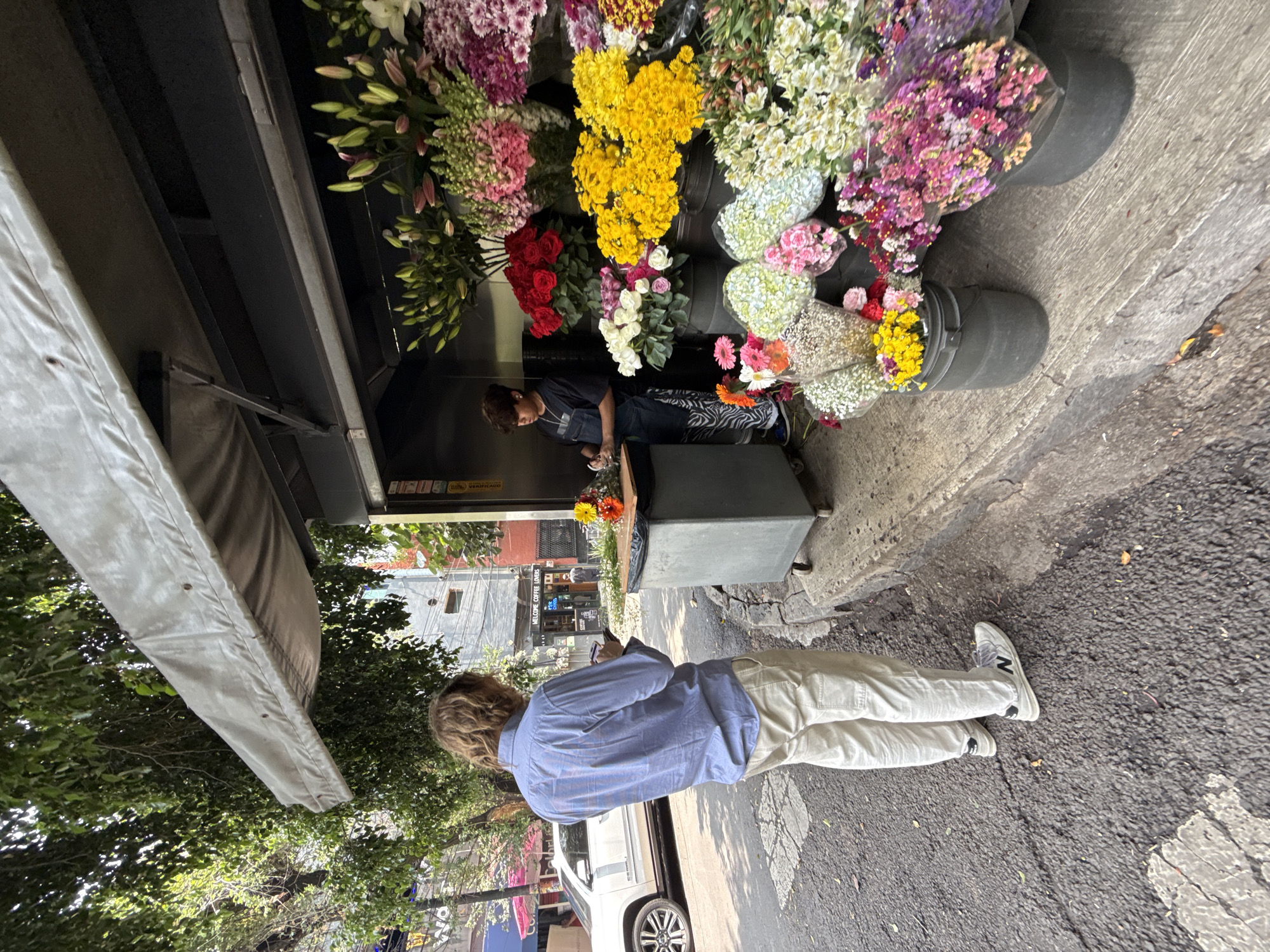 The image captures a street scene in Mexico City, Mexico. A man with blond hair, wearing a blue collared shirt and khaki pants, stands on the sidewalk, looking at his phone. He faces a flower stand displaying numerous buckets of colorful flowers, including lilies, roses, and chrysanthemums. Behind the flower stand, a woman wearing a mask is tending to the flowers. The flower stand is positioned in front of a building with an awning. In the background, a white car is partially visible, and a glimpse of street life can be seen, including buildings and trees. The street surface appears to be asphalt with visible cracks and markings.