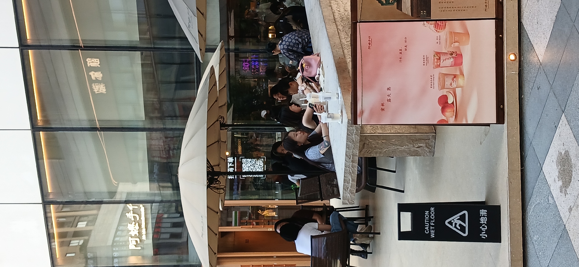 In the Jiulongpo District of China, an outdoor seating area of a cafe is depicted. Several people are seated at a large, stone-topped table under a light-colored umbrella. They appear to be friends or acquaintances, with some engaged in conversation and others looking at their phones. The cafe has large glass windows and a modern architectural design. A "Caution Wet Floor" sign is visible in the foreground, indicating a potential safety hazard. A pink poster displaying images of fruit and beverages is also present near the seating area. The overall scene suggests a casual social gathering at a contemporary urban establishment.