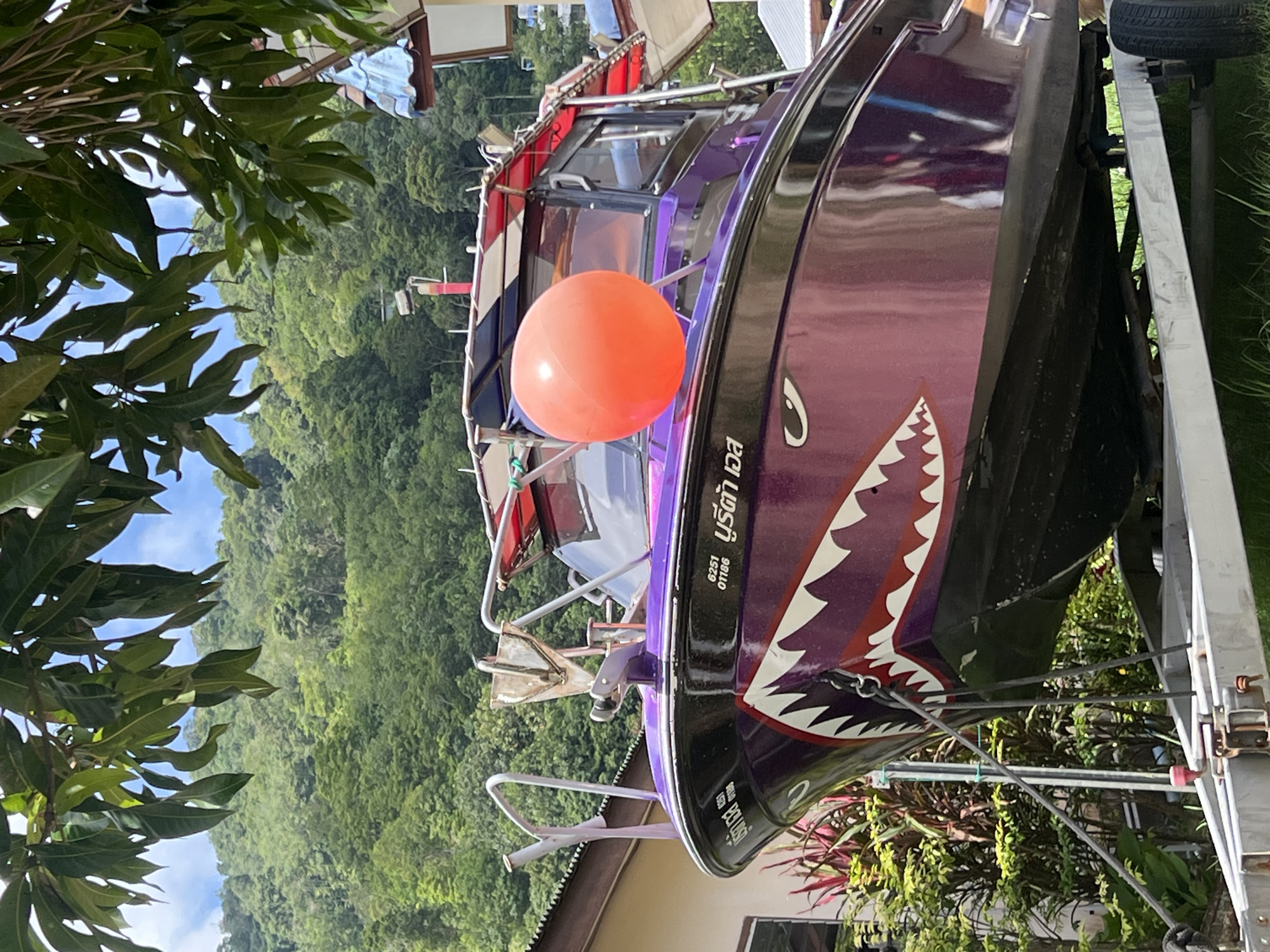 A purple speedboat with a shark mouth decal on its bow is parked on a trailer on a grassy area in front of a building. The boat has a red and white striped canopy. A large orange buoy is attached to the side of the boat. In the background, a lush green hillside covered in trees rises under a blue sky with scattered clouds. The scene appears to be in a tropical location, likely Choeng Thale, Thailand, given the vegetation and architecture. No people are visible in the image.