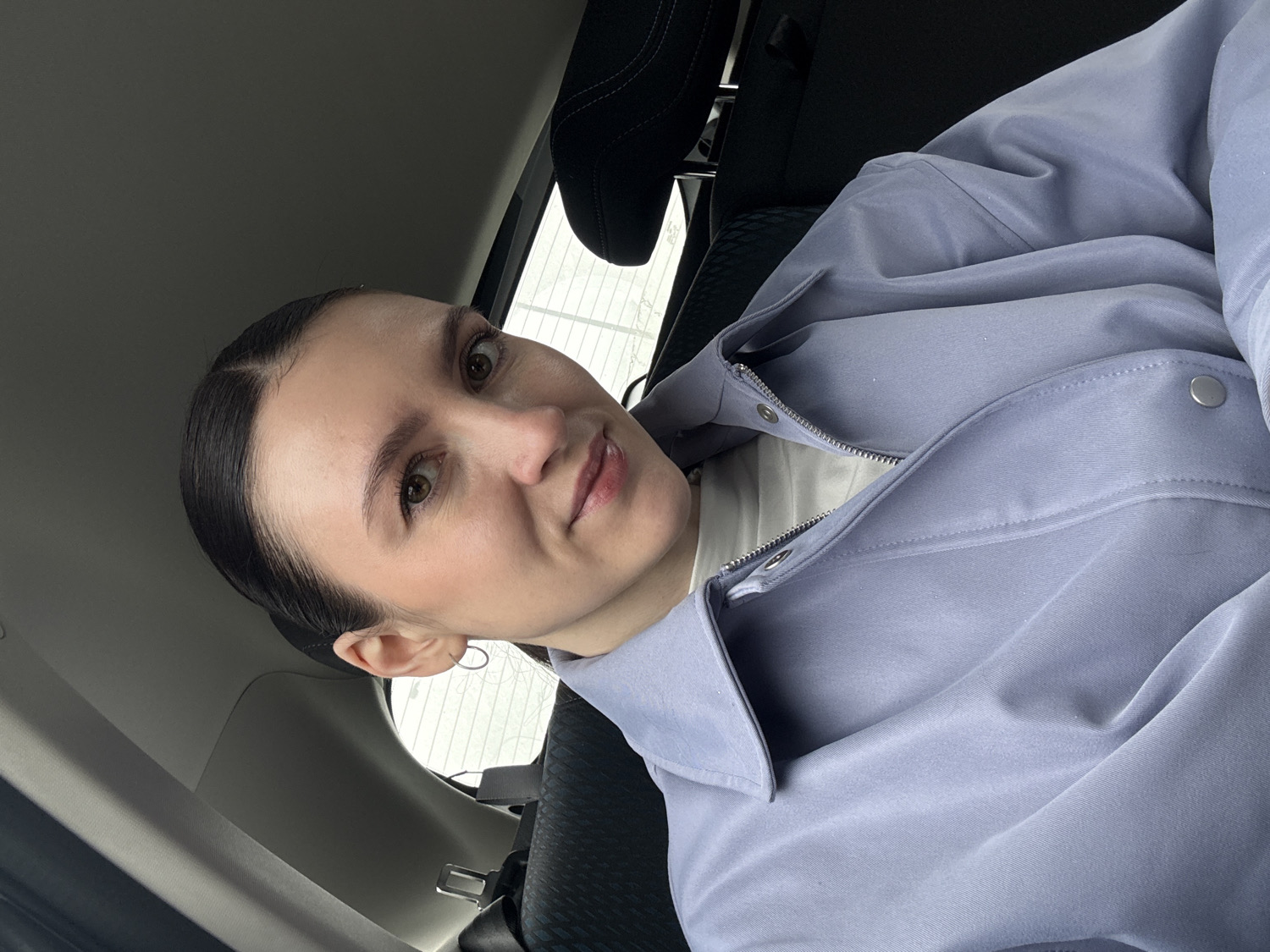 A woman is depicted in a medium-close-up, seated inside a car in Kazan, Russia. She is positioned towards the left of the frame, looking directly at the camera with a neutral to slightly pleasant expression, her lips gently parted. Her dark hair is pulled back neatly from her face, revealing a distinct part. She has light-colored eyes and appears to be wearing minimal makeup, including subtle eyeliner and lip color.
She is dressed in a light blue or periwinkle-colored jacket with a collar, visible zipper, and silver snap buttons. A white shirt is visible underneath the jacket. The car's interior is visible around her, including a dark headrest behind her and a car seat. Through the car window in the background, a soft, diffused light suggests it is daytime, possibly an overcast or cloudy day. No specific outdoor elements are clearly discernible beyond the light, indicating a general outdoor setting. A small, subtle hoop earring is visible in her left ear. There is no visible text in the image.