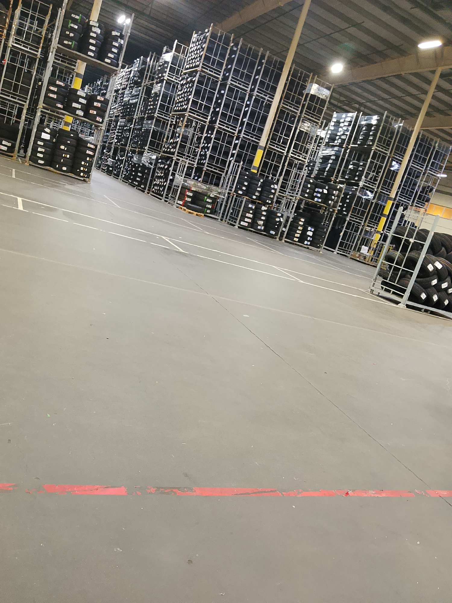 An interior view of a warehouse in Elektrougli, Russia, shows rows of tall shelving units filled with stacks of tires. The tires are black and appear to be new, some are wrapped in plastic. The warehouse floor is a smooth, gray concrete surface with white and red painted lines. The ceiling is high and industrial, with visible support beams and lighting fixtures. The overall impression is of a large-scale storage facility for automotive tires.