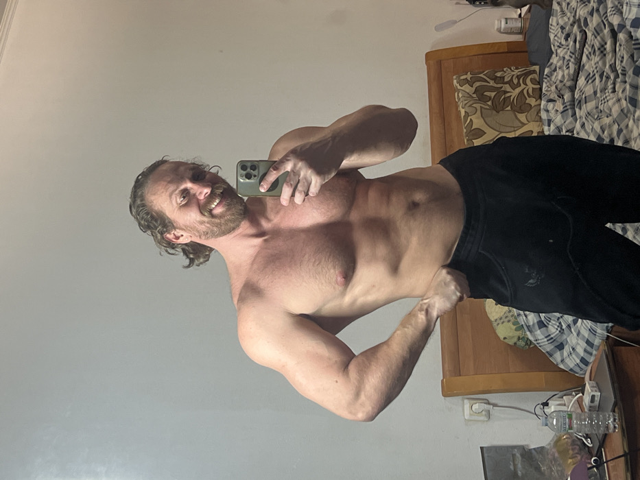 An adult male with a beard and wavy brown hair is depicted shirtless from the waist up, showcasing a muscular physique. He is smiling and holding a dark-colored smartphone in his right hand, reflecting his image in a mirror selfie. His left hand rests on his left hip, slightly pulling down his dark-colored athletic pants. The scene is an indoor setting within Saint Petersburg, Russia. The background consists of a plain, light-colored wall. To the right, a wooden headboard and a bed covered with a blue and white plaid duvet are visible, along with a brown and beige patterned pillow. To the left, a wooden nightstand holds a plastic water bottle with a "Bulgaria" label, a white rectangular object, and a partially visible laptop or tablet. An electrical outlet is present on the wall above the nightstand.