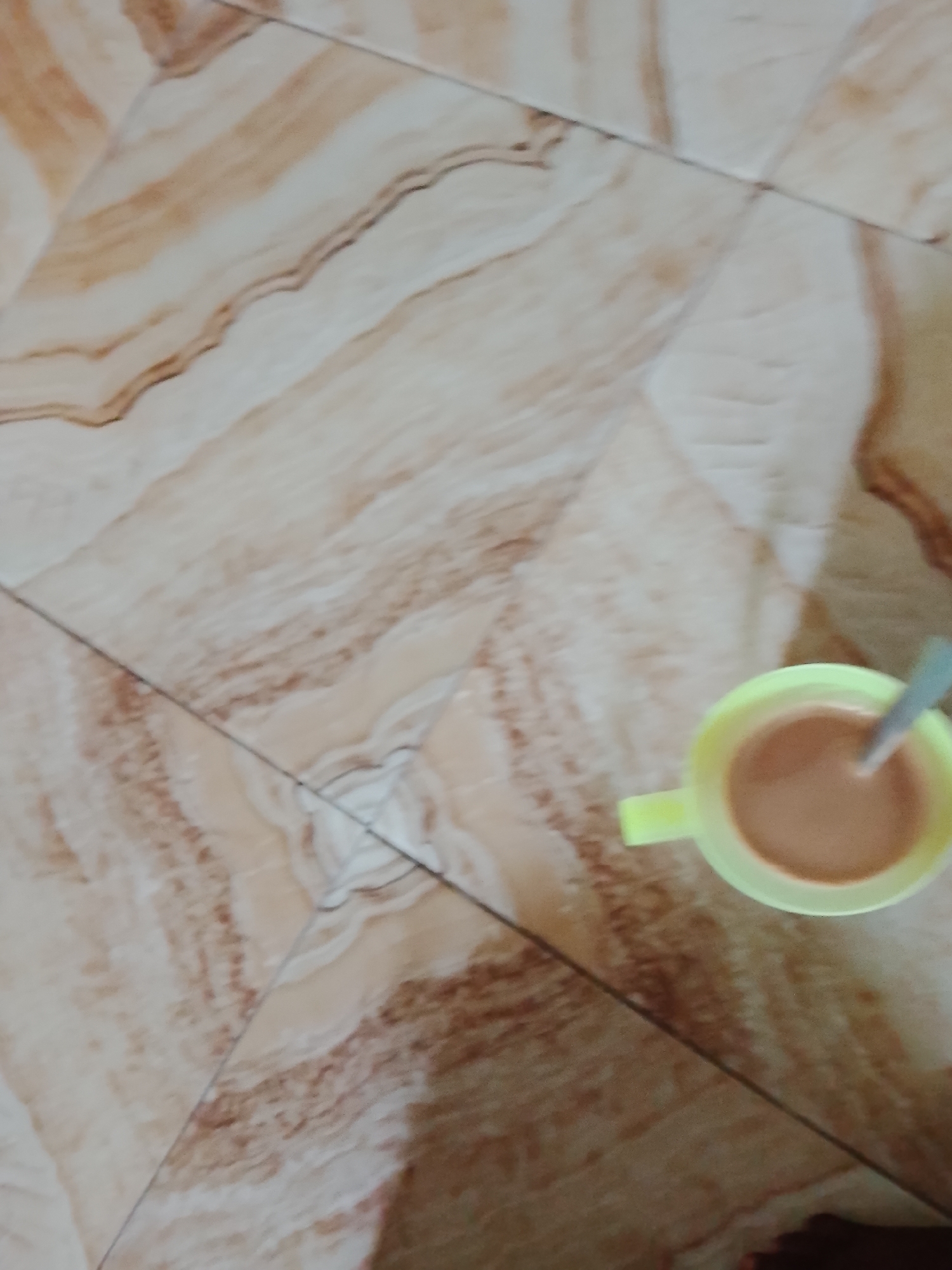 An overhead shot shows a pale yellow mug with a light brown liquid and a spoon inside, resting on a tiled floor. The floor tiles are a light beige with brown marbling. The scene is lit from above. The image appears to have been taken in a domestic setting, possibly a kitchen or living area, in Pantinapu, Nigeria. The image does not provide any specific details to confirm the location.