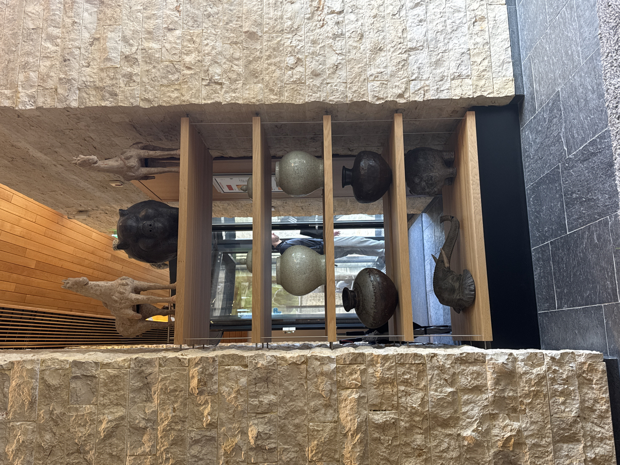 The image shows an indoor art installation. The installation is framed by light-colored stone walls above and below, and wooden beams forming vertical dividers. Within these dividers, a variety of vase-like objects and sculptures are displayed. The vases are of different sizes, shapes, and colors, some light and some dark. Sculptures appear to include figures resembling animal forms. Reflected in the glass behind the display is a person looking into the installation. The location is Noordwijk, Netherlands.