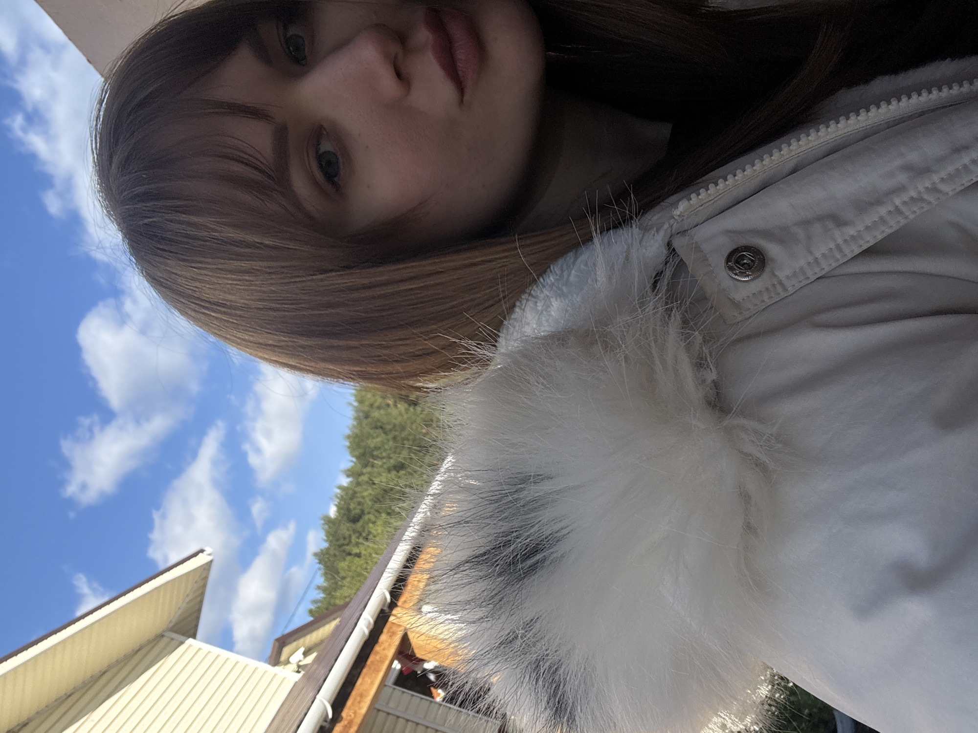 A photograph appears to have been taken by a person facing upward and toward their left side, capturing a view of their face, the upper portion of a white jacket with a furry trim, and a background featuring a building, trees, and a blue sky with clouds. The person has light skin, brown hair with bangs, and neutral-toned lips. The jacket is light-colored with a button visible. In the background, a building with a light yellow exterior and a brown roof is partially visible, along with a patch of green trees and a bright blue sky dotted with white clouds. This scene is likely located in Chaikovsky, Russia.