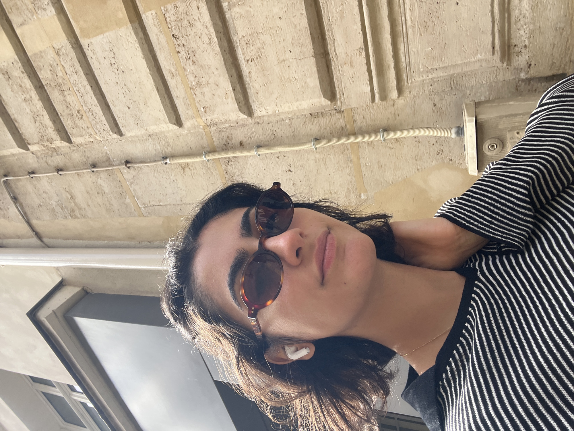 A low-angle selfie captures a young woman looking upwards towards the camera. She has dark, shoulder-length hair and is wearing round, tortoiseshell-patterned sunglasses. A white AirPods Pro is visible in her right ear. Her expression appears calm or neutral, with her lips slightly parted. She is dressed in a black and white striped long-sleeved top, and a thin gold chain necklace can be seen resting on her collarbone.
The background features an old, light-colored stone or concrete ceiling with prominent horizontal grooves or beams. The surface exhibits texture and some signs of age. A white electrical conduit runs horizontally across the upper part of the background, secured by clips, leading to a light-colored rectangular electrical box mounted on the wall or ceiling on the right side of the frame. On the far left, a dark-framed window or architectural detail with geometric patterns is partially visible.
The lighting is bright and sunny, suggesting it is daytime, with clear highlights on the woman's hair and the architectural elements. The setting evokes an urban environment, consistent with the historical architecture found in Paris, France. No visible text is present in the image.
