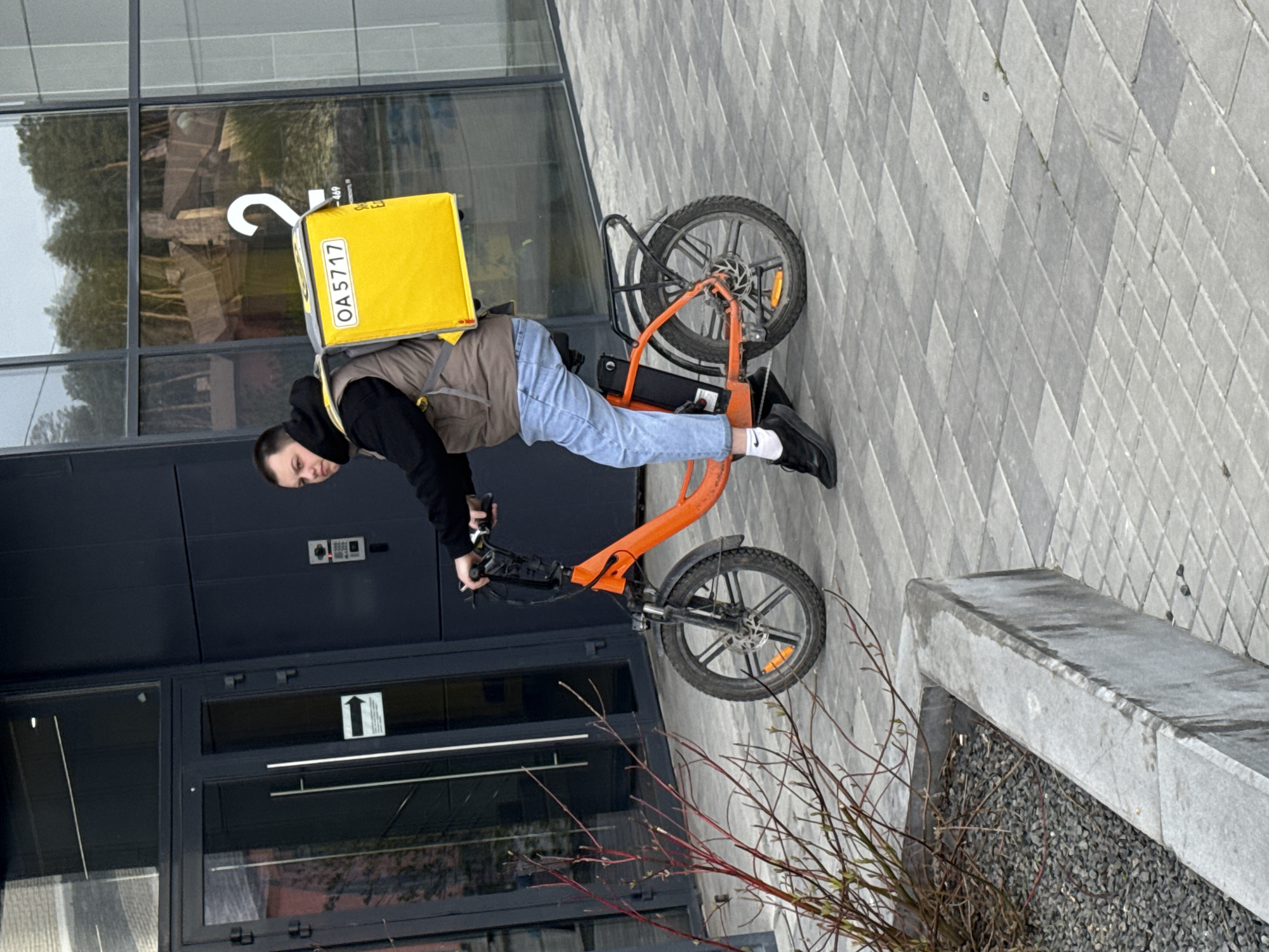 A delivery rider stands beside an orange electric bicycle or scooter in an urban environment in Yekaterinburg, Russia. The person, appearing to be a young male, wears a black hoodie, a brown vest, blue jeans, white socks, and black shoes. A large, bright yellow insulated delivery box, possibly a backpack, is strapped to their back. A white label on the box displays the alphanumeric text "OA5717". A white, hook-shaped or question mark-like symbol is also partially visible on the top of the box.
The rider stands with their hands on the handlebars of the orange bike, which features small wheels and a rear rack. The setting appears to be outside a modern building with dark grey architectural panels and large reflective glass windows. Reflections of trees and possibly other structures are visible in the windows. A keypad or intercom system is mounted on a dark panel next to a door. The ground is paved with grey interlocking blocks arranged in a diagonal pattern. To the right, a low concrete planter box filled with dark gravel contains sparse, bare reddish-brown branches. The lighting suggests it is daytime, possibly overcast, with no harsh shadows. The rider's expression is neutral as they look towards the camera.