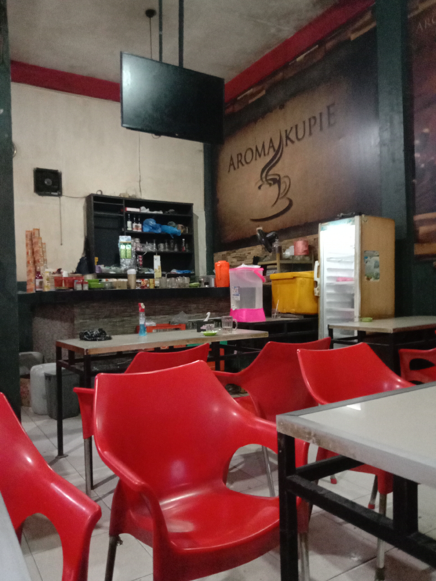 The image depicts the interior of a casual cafe or warung in Lhokseumawe, Indonesia, likely in the evening or at night, given the reliance on artificial lighting. The establishment appears to be empty of customers.
In the foreground and midground, multiple bright red plastic chairs are arranged around simple rectangular tables with light-colored surfaces and dark metal frames. The floor is covered with light-colored tiles.
Towards the back, a long service counter runs across the left side of the frame. Behind the counter, shelves are stocked with various bottles, plastic containers, and what appears to be beverage-making equipment. Above the counter, a black shelving unit holds more items. A small, black wall-mounted speaker or fan is visible on the wall to the left, and a flat-screen television is mounted high above the counter.
On the right side of the image, a light-colored refrigerator with an open door stands next to a yellow cooler and a pink and clear water dispenser. A prominent graphic on the wall behind these items features the text "AROMA KUPIE" in a stylized font, accompanied by a coffee cup illustration. A horizontal red beam runs along the top of the wall, contrasting with the off-white wall color. The overall impression is that of a functional, unpretentious eatery, seemingly specializing in coffee, as suggested by its name.