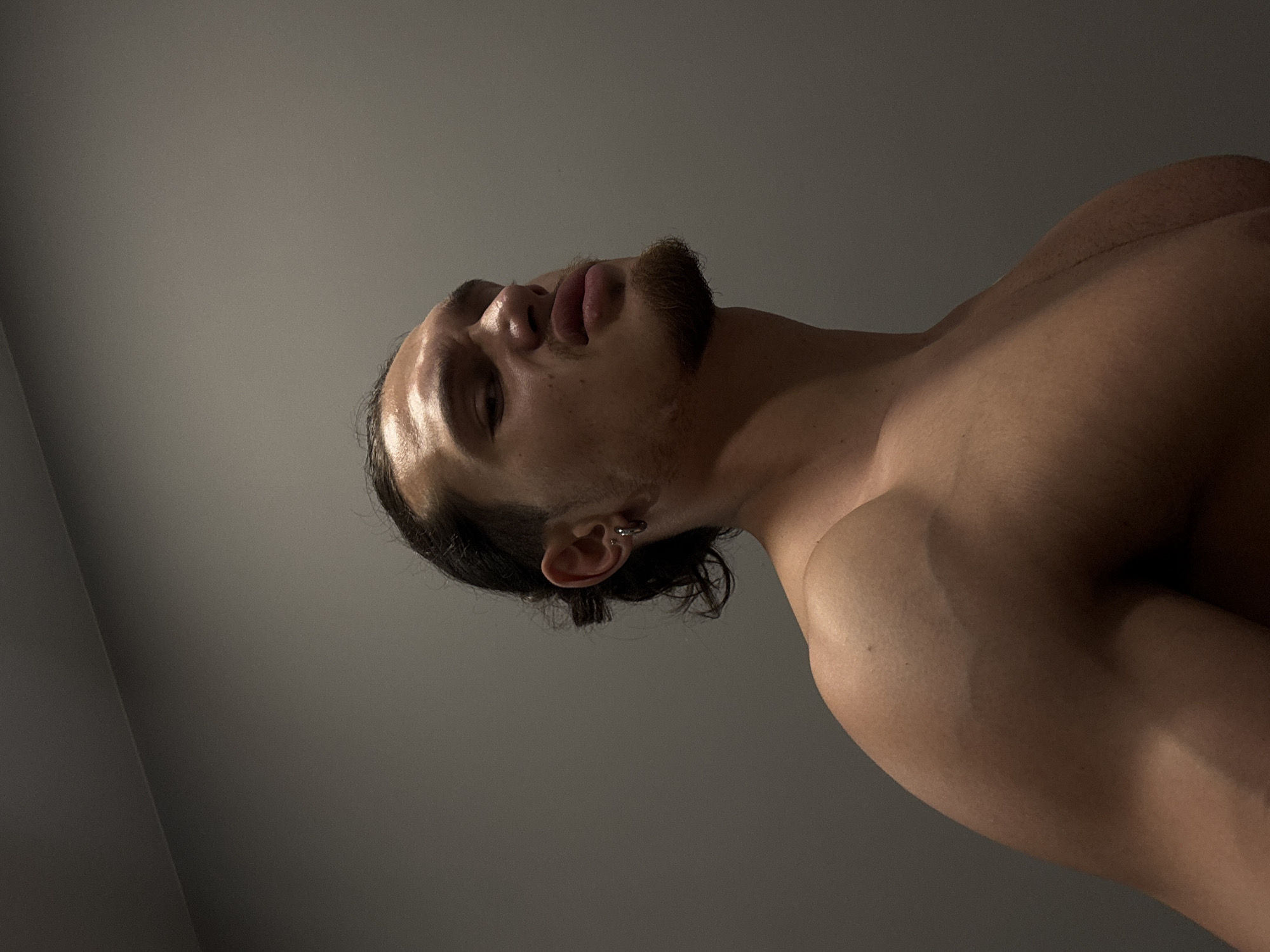 The image is a low-angle shot of a shirtless man with dark hair pulled back, a beard, and an earring in his left ear. His head is tilted back, and his face is illuminated by a light source. His shoulders and chest are visible. The background is a plain grey surface. No details indicate the location is in Sofia, Bulgaria.