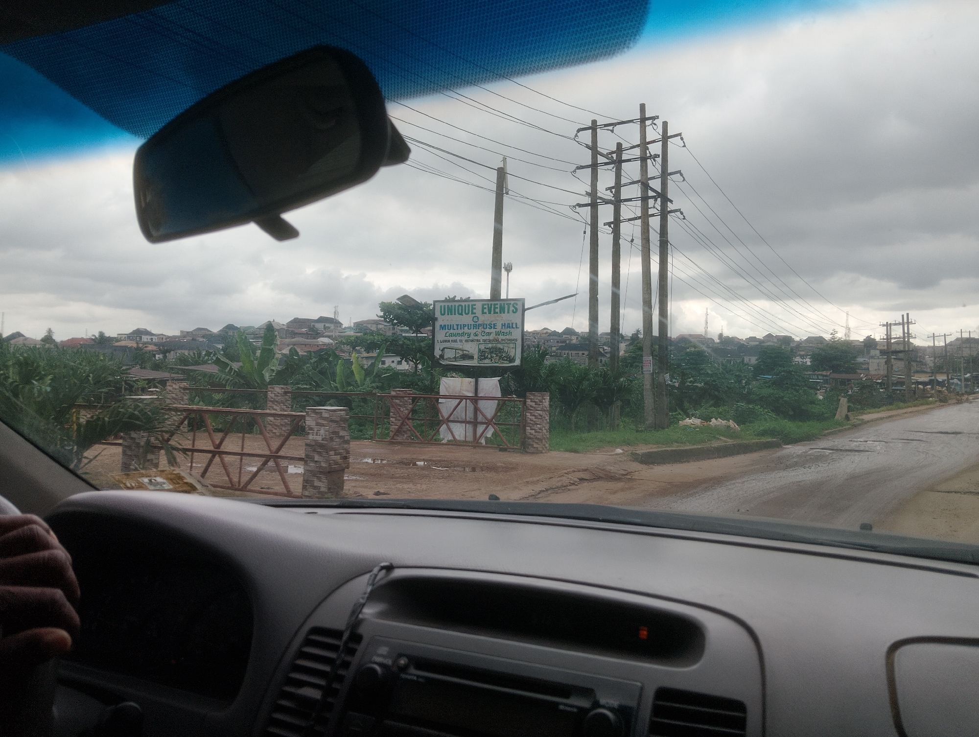 This media file is a first-person perspective shot from inside a car, looking out through the windshield. The view captures a street scene in Iju, Nigeria, under an overcast sky, suggesting it might be a cloudy day, possibly in the late morning or early afternoon.
The foreground is dominated by the car's dashboard and steering wheel, with a driver's hand visible on the wheel. The interior is dark, with some instruments dimly lit.
The view outside the car shows a somewhat rural or semi-urban setting. There are houses with varied roofing styles in the distance, nestled amongst lush green vegetation, likely trees and banana plants. Prominent in the mid-ground are several tall utility poles with numerous power lines crisscrossing the sky. Adjacent to the road, there is a fenced entrance made of brick pillars and metalwork.
A sign is clearly visible, mounted on a pole. The text on the sign reads: "UNIQUE EVENTS & MULTIPURPOSE HALL Laundry & Car Wash" followed by an address and phone numbers: "LAJUWURA, TEL: 05074607958, 09013848284, 0073469435". This indicates the presence of a local business offering event services and other facilities. The road itself appears to be paved but shows signs of wear and tear, with several potholes. The overall impression is of a typical day in a Nigerian town, with everyday infrastructure and local commerce visible.