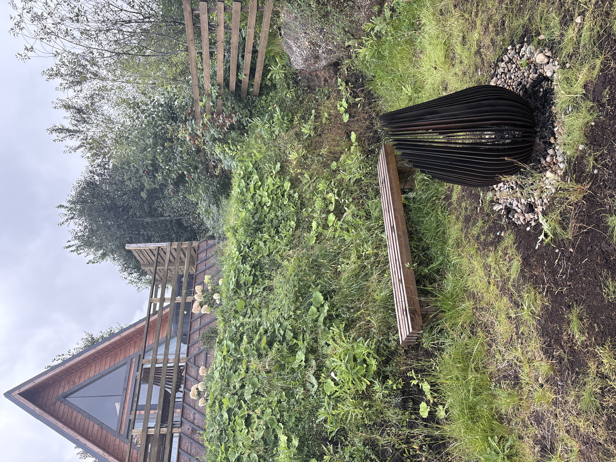 The image depicts a fire pit, a wooden bench, and a steep, overgrown embankment with a modern, triangular-shaped wooden house in the background. The fire pit is black, tear-drop shaped, and made of vertically arranged metal slats, surrounded by small grey and white stones and dark soil. The wooden bench is located to the left of the fire pit and is made of several parallel wooden planks. The embankment is covered with lush green vegetation, including large-leafed plants and tall grasses. The house is a prominent feature, with a large triangular window and wooden railings on a balcony. A wooden fence runs horizontally across the upper right portion of the image, bordering the overgrown area. The sky is overcast. The scene appears to be located in a natural, possibly rural or suburban, setting outside of a populated area. There are no people present in the image.
