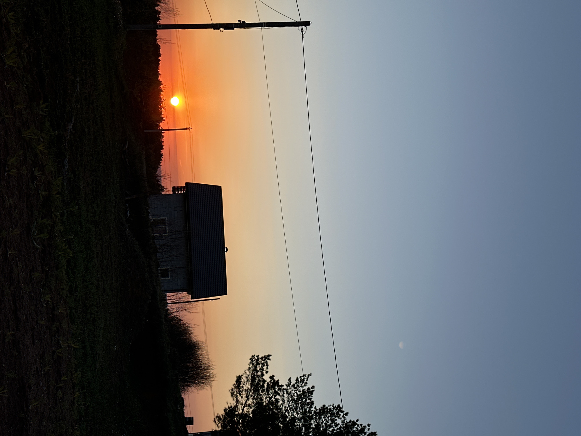 Captured in Прикамский, Russia, the photograph presents a landscape at what appears to be either sunrise or sunset. The sun, visible in the upper left, casts an orange glow across the sky. A dark, silhouetted house stands in the midground, adjacent to a field and some trees. Power lines cut across the sky, and a faint moon is visible in the upper right.